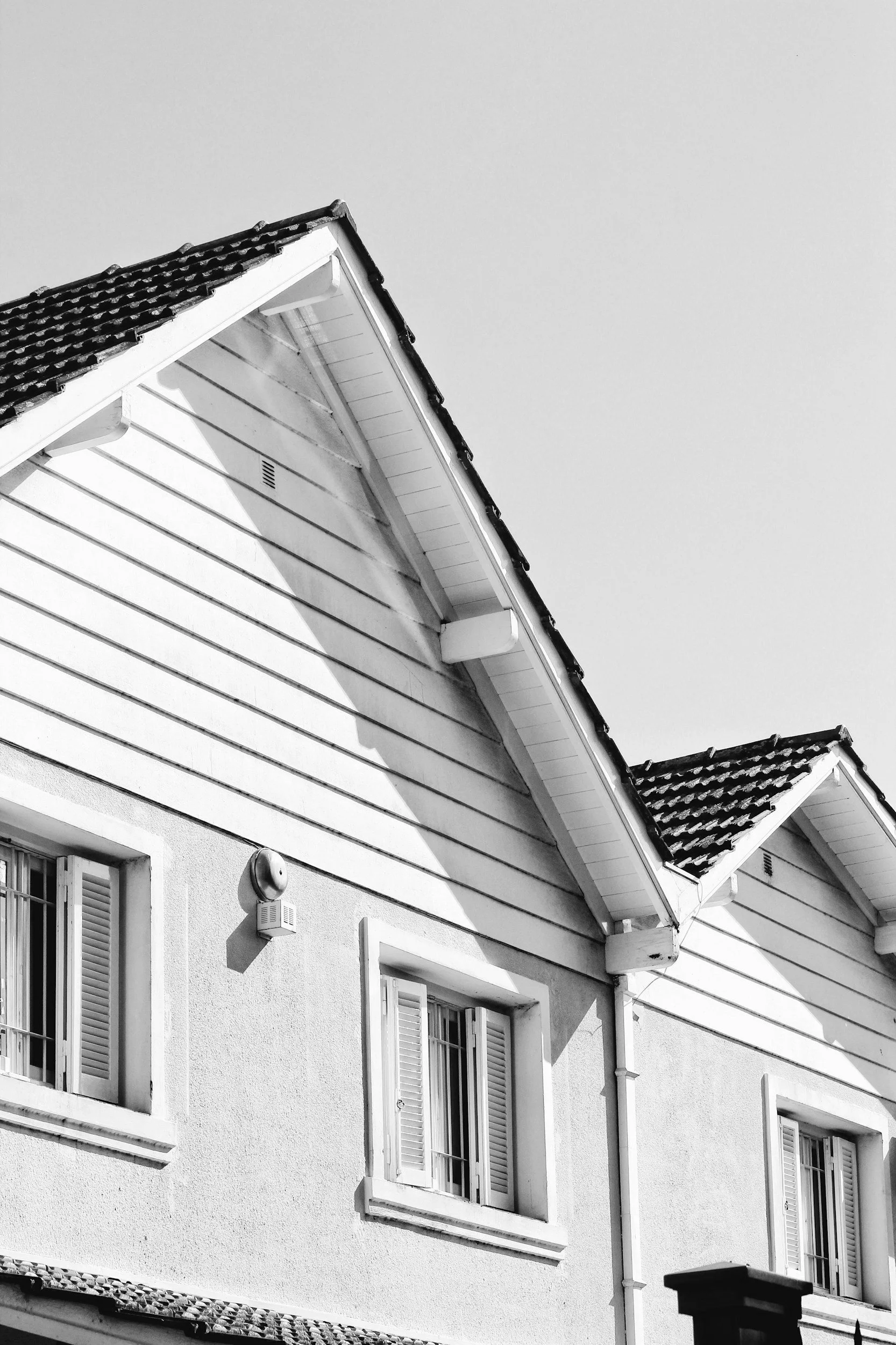 Black and white photo of the upper part of a residential house showing its gable roof, windows with shutters, and exterior wall.