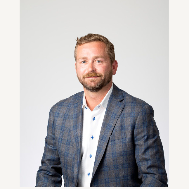 A man with a beard and short hair, wearing a plaid blazer and a white shirt, smiling against a plain background.