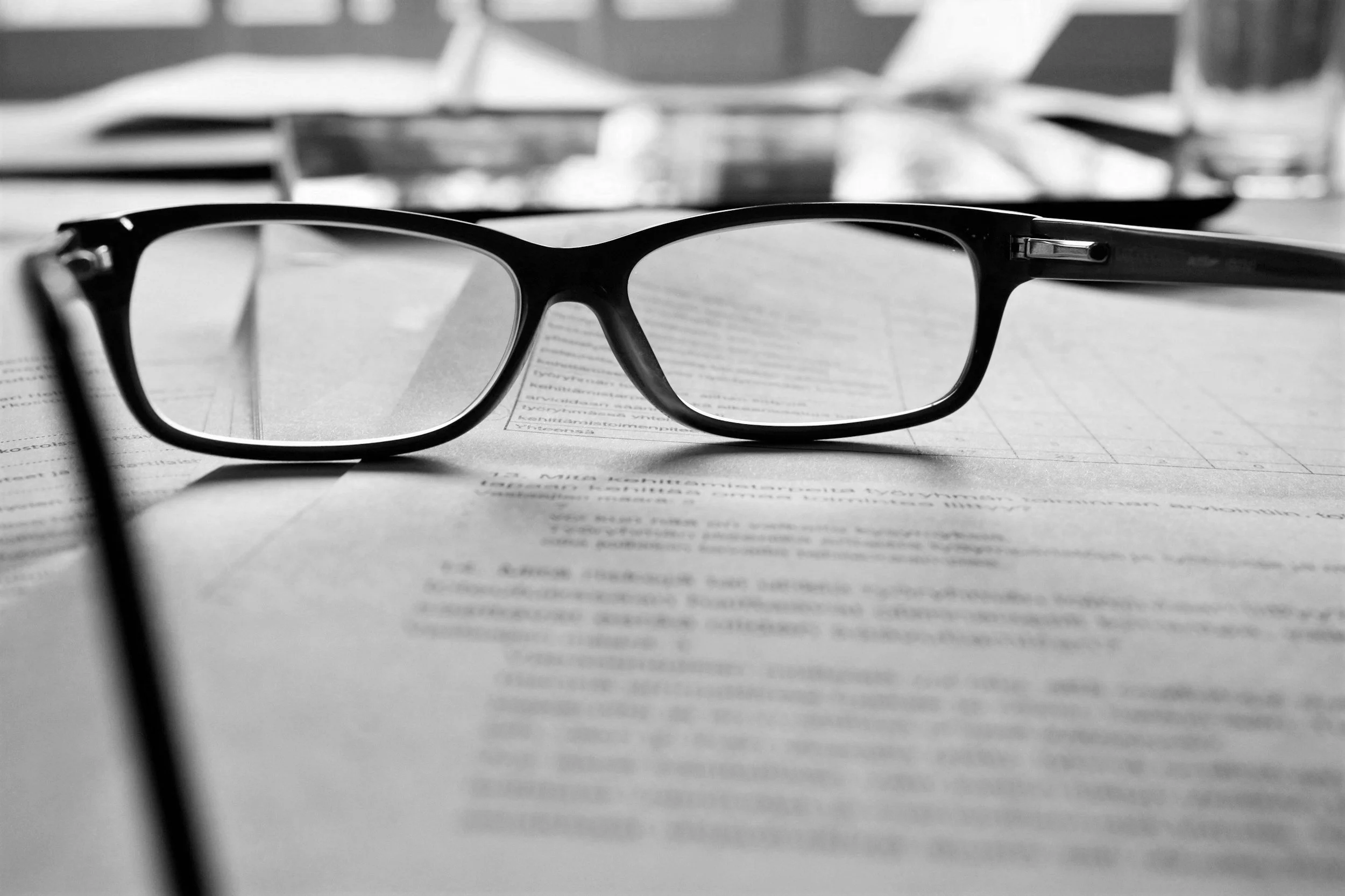 A pair of black eyeglasses resting on a newspaper or printed document on a table, with a blurred background.