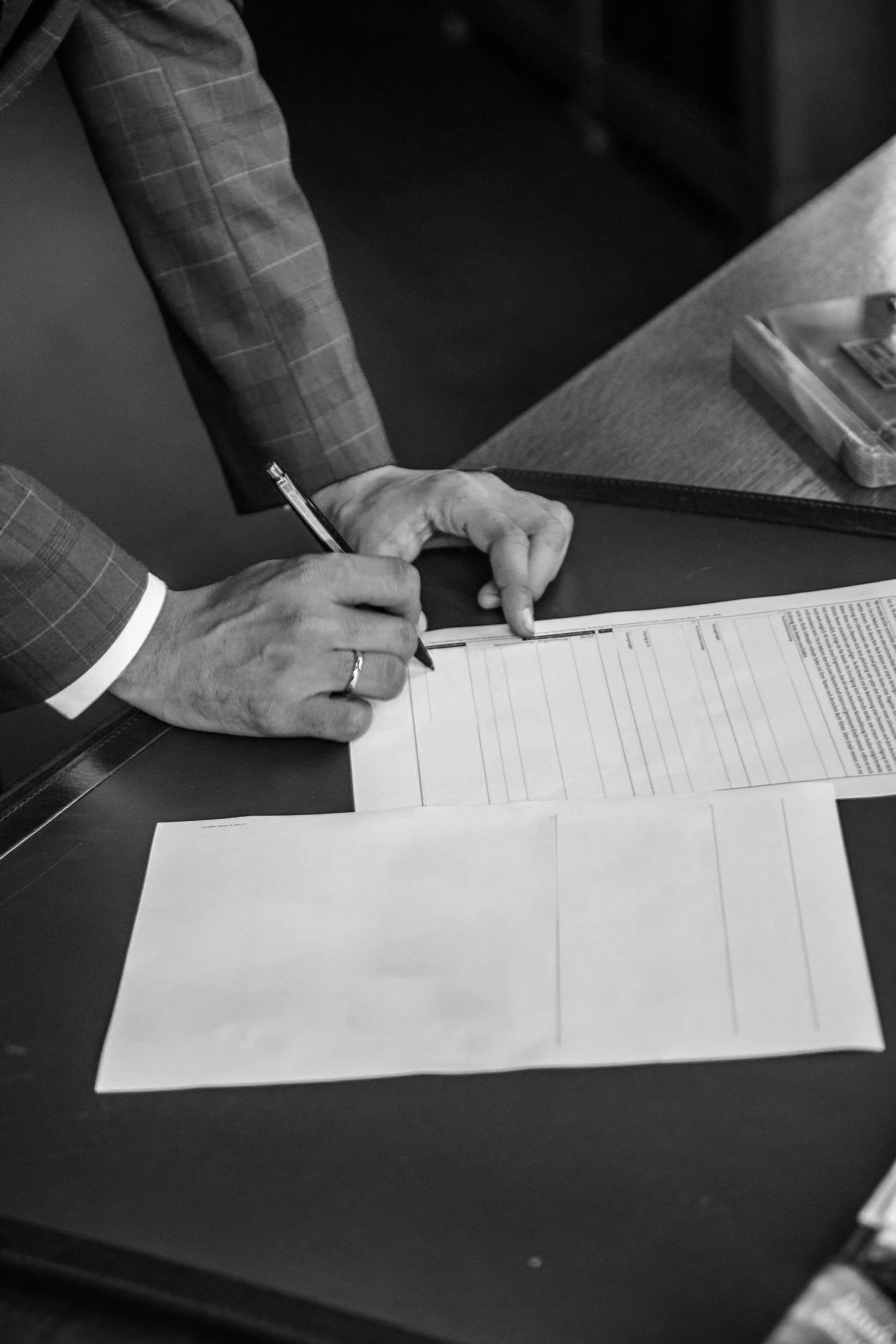 A person in a checked suit is writing on a paper with a pen while placing their other hand on the paper. There are documents and a ruler on the desk.