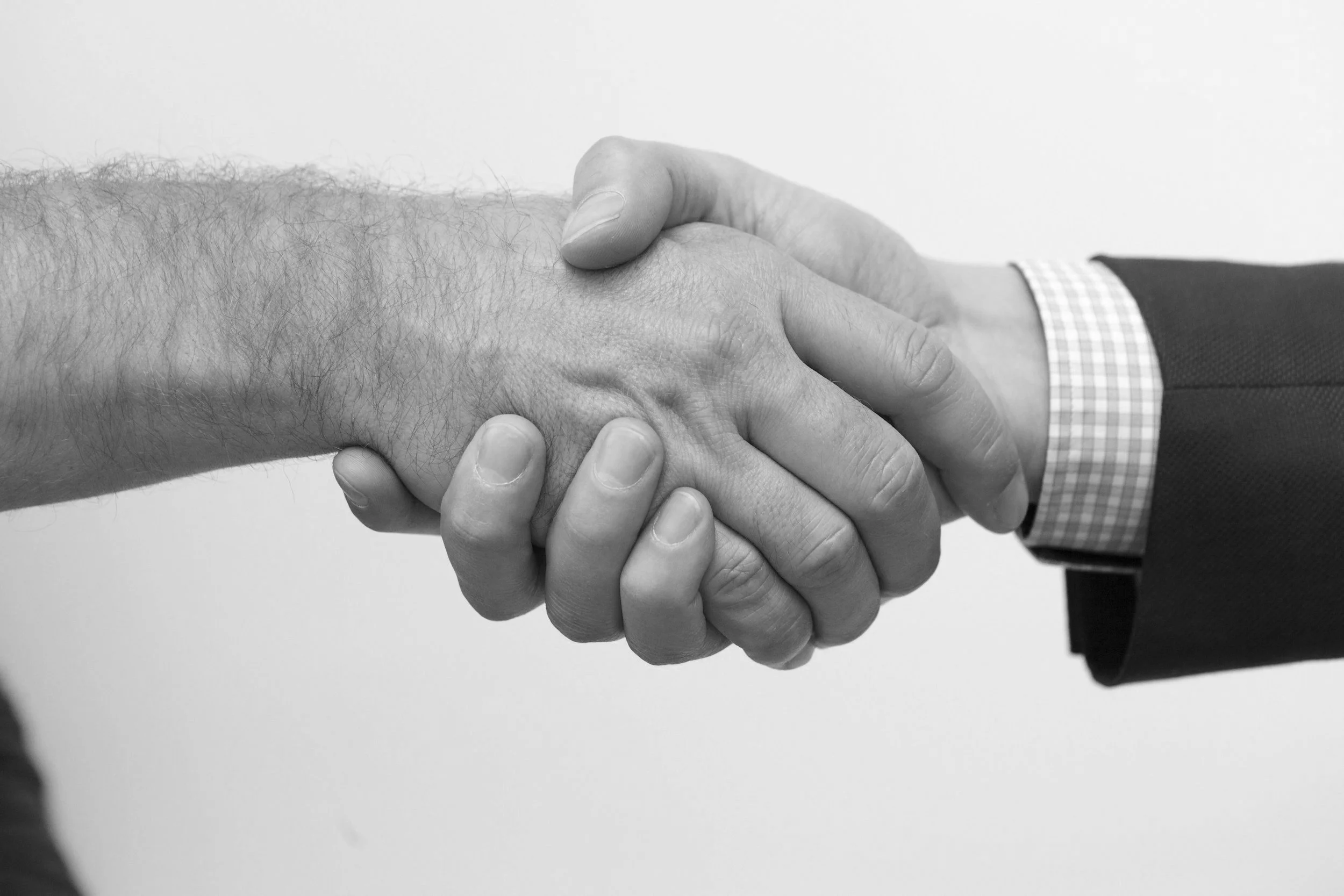 Two people shaking hands, one with a hairy arm and the other wearing a suit jacket and a checkered shirt, black and white photo.