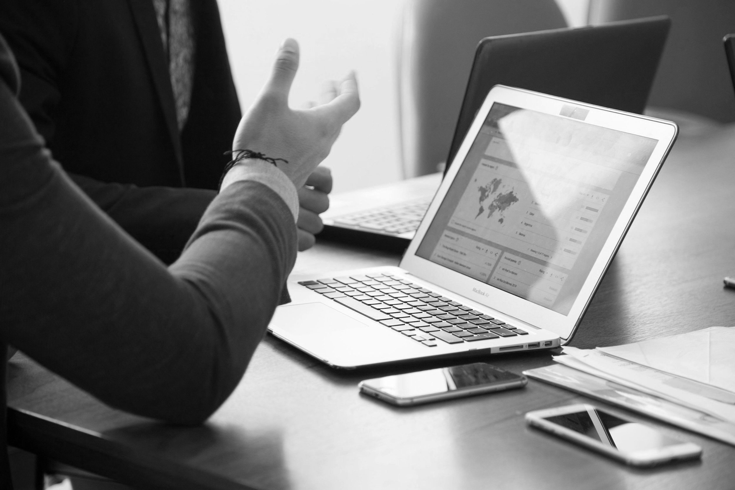 A person gesturing while sitting at a desk with a MacBook laptop, a phone, and documents, during a meeting or discussion.