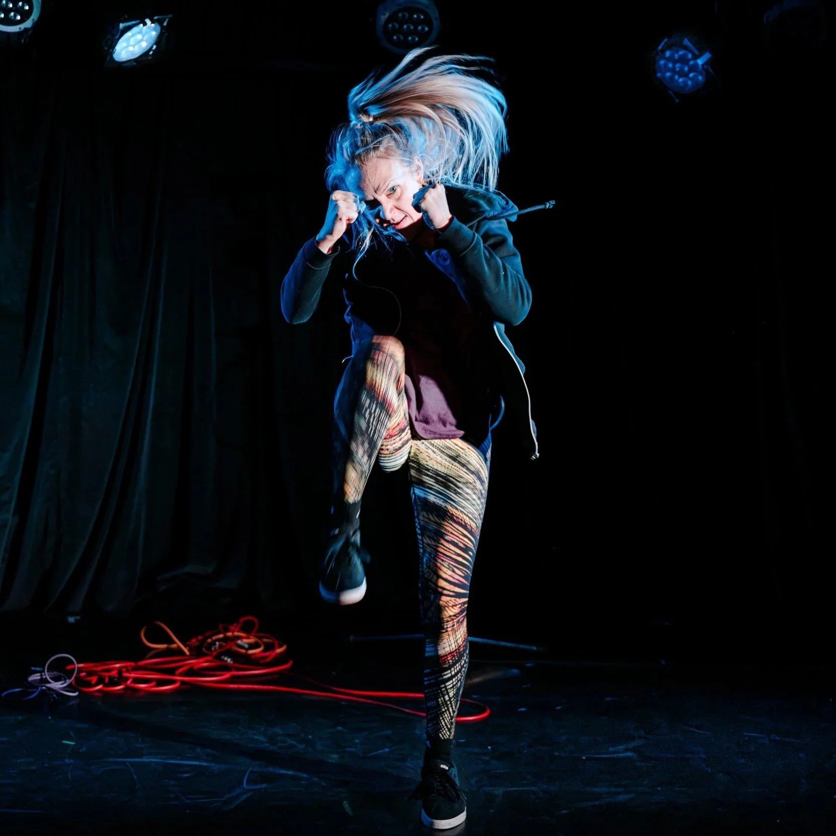 Production image. A woman kickboxing aggressively on a dark stage