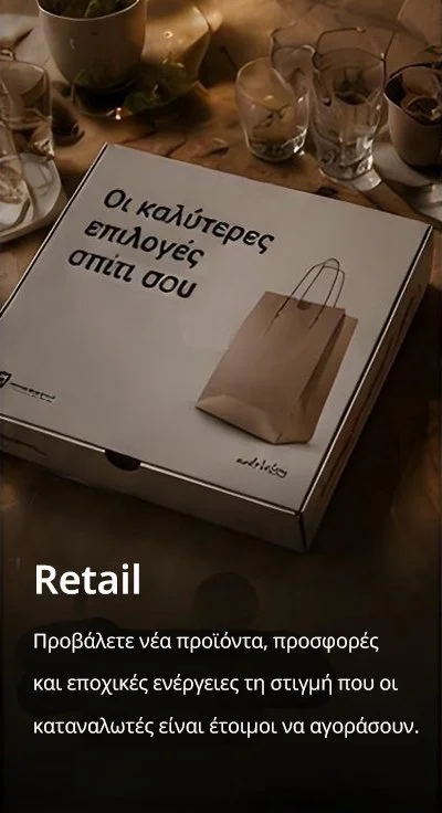 A pizza box package with Greek text and a paper shopping bag illustration on it, surrounded by glasses and cups on a wooden surface.