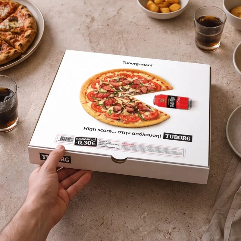 A person holding a Tuborg pizza box with a picture of a pizza topped with pepperoni, green peppers, and onions, surrounded by soda glasses and other food items on a table.
