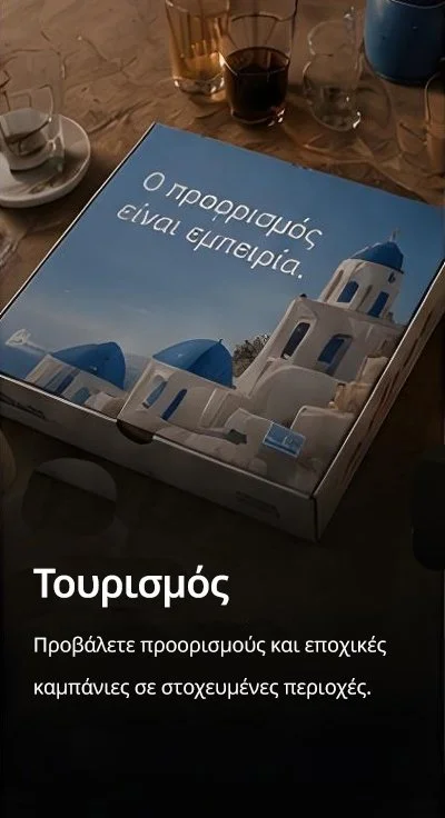 A pizza box ad with Greek text and visual of white buildings with blue domes, resembling Santorini, Greece, on a table with drinks and a cup nearby.
