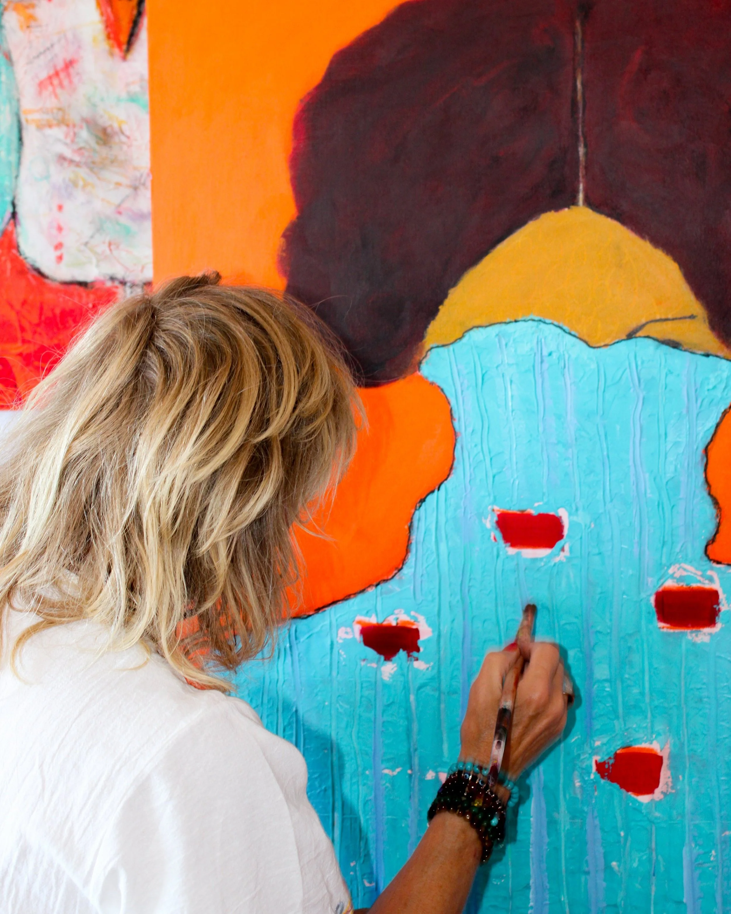 An artist with long blonde hair painting an abstract colorful mural on a wall.