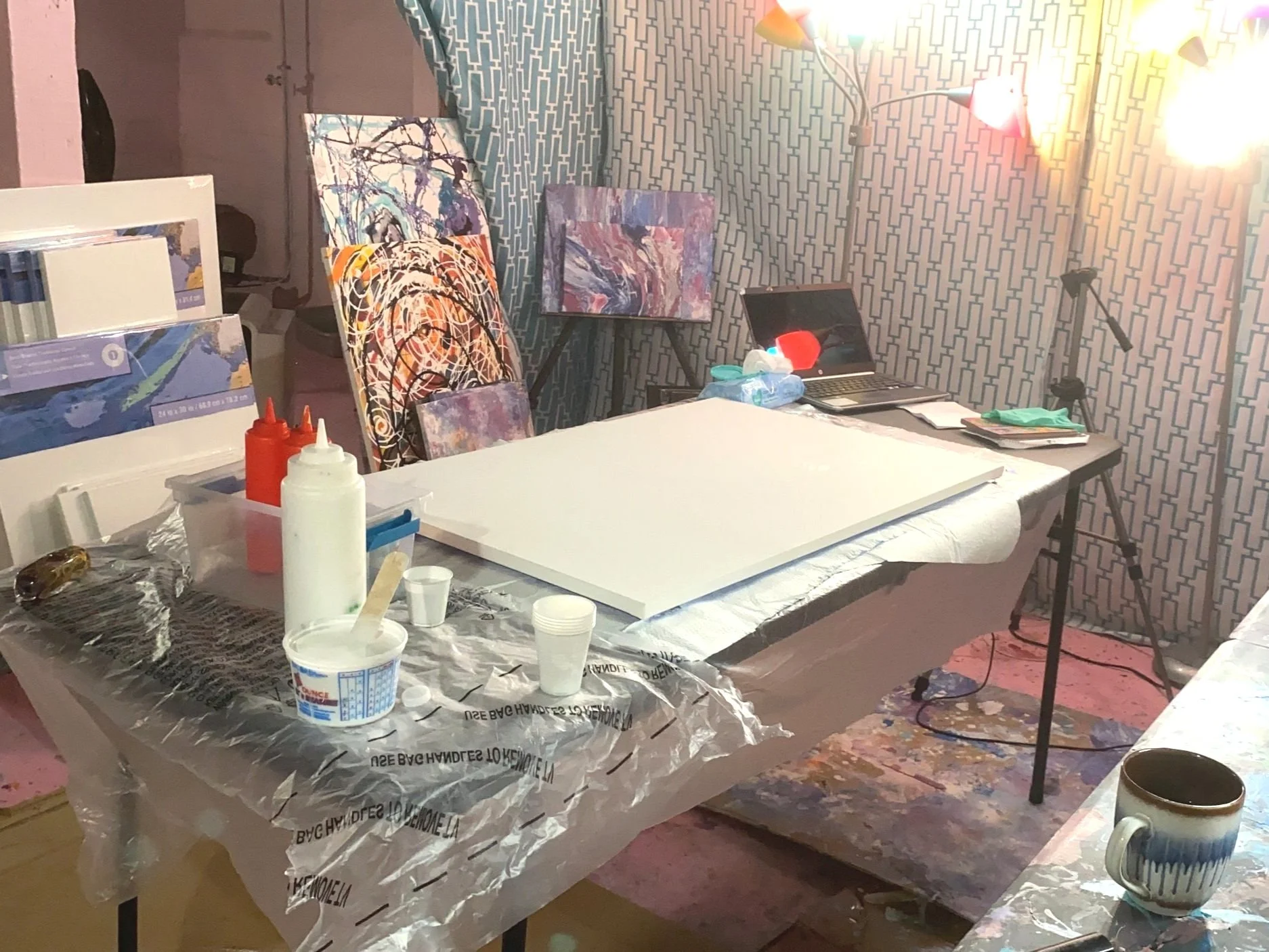 Interior of an artist's studio with a work table covered in plastic and art supplies, including paint bottles, a cup with a stirring paddle, and artworks in progress displayed against a patterned wall. A laptop and various art books are also on the t
