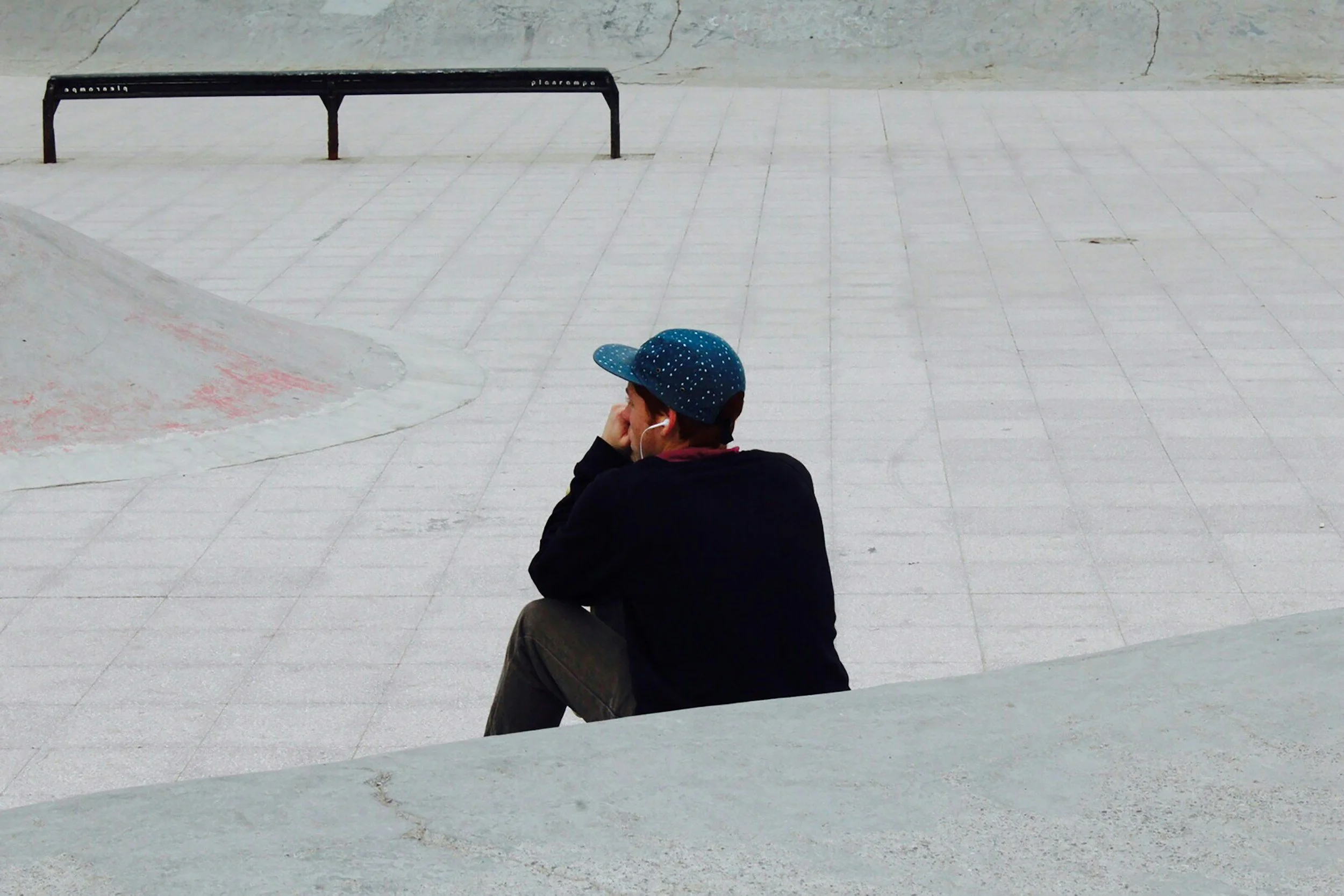 A person wearing a blue cap with white dots, black jacket, and face mask sitting alone on a concrete surface, looking away in an empty outdoor area.