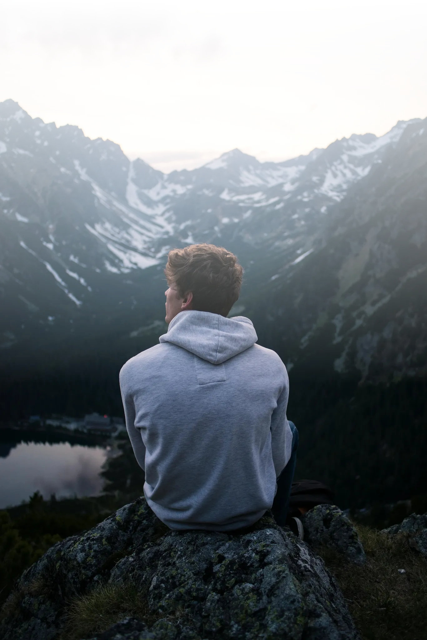 A person sitting on a rock, facing a mountain range with snow patches, overlooking a lake during dusk or dawn.