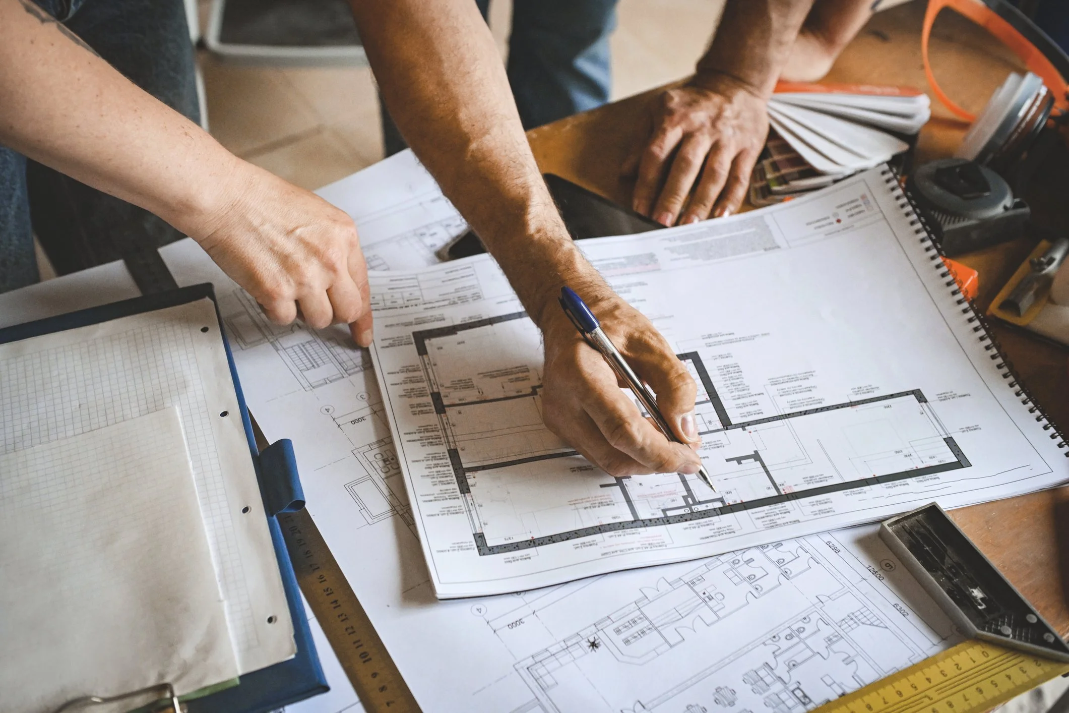 People reviewing architectural blueprints on a wooden table, with various tools and notebooks nearby.