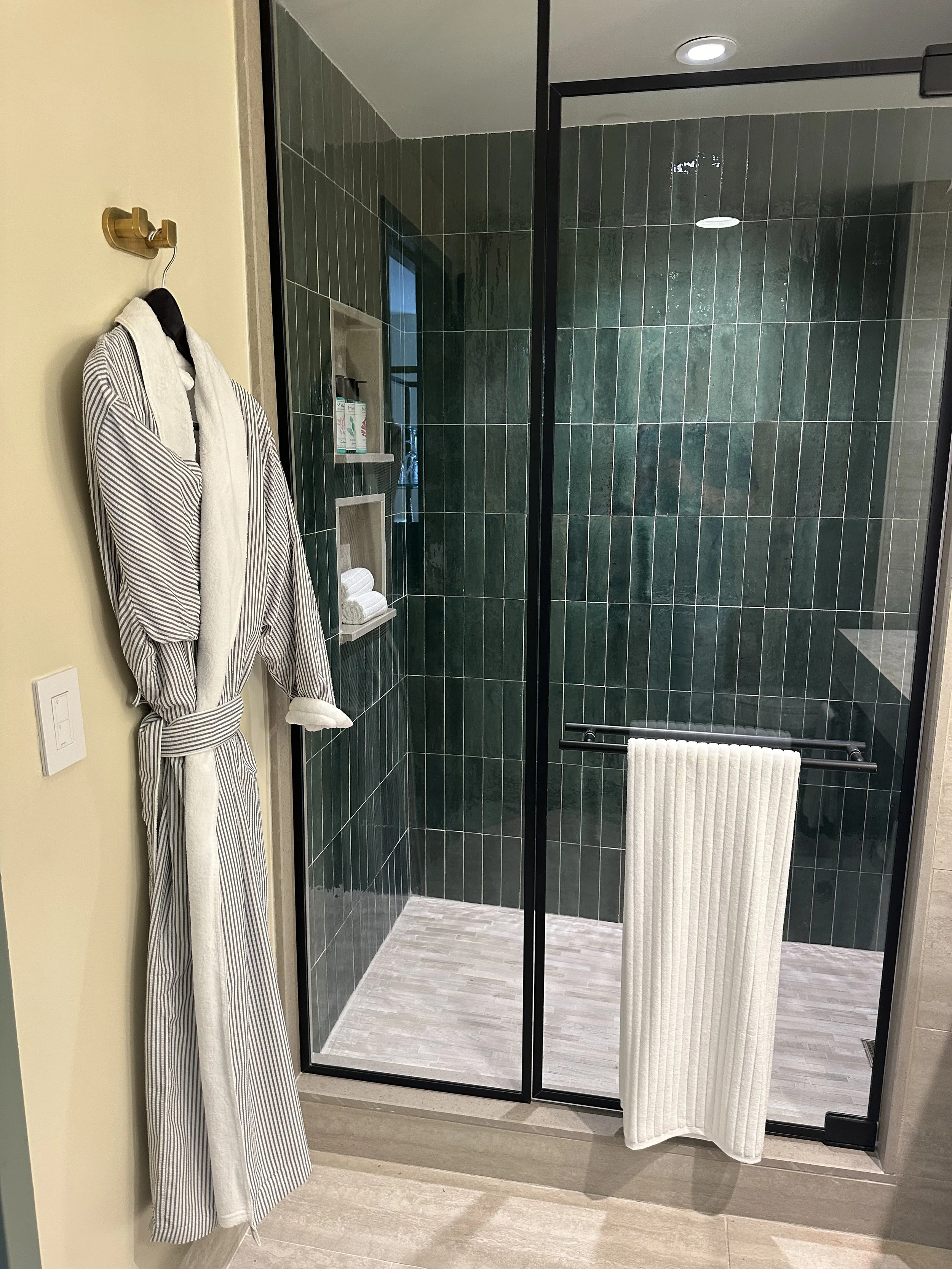 A glass-enclosed shower with dark green tiled walls, a built-in shelf with shampoo and soap, a white towel hanging on a towel bar, and a robe hanging on a hook outside the shower.