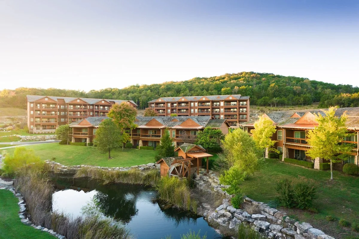 Residential buildings with multiple floors and balconies, surrounded by greenery, near a pond with rocks, and a hilly landscape in the background.