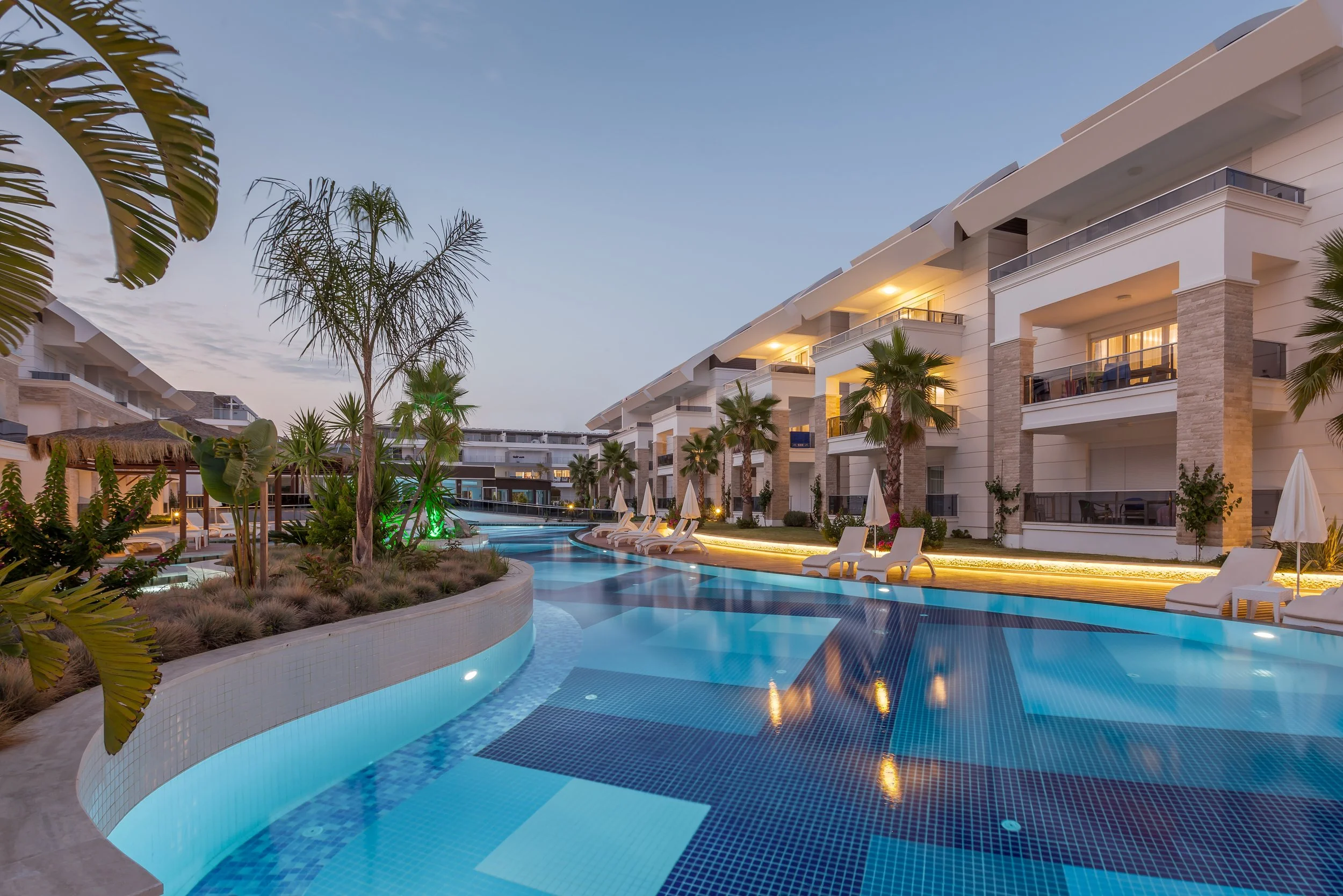 Resort pool area with lounge chairs, palm trees, and modern white apartment buildings at dusk