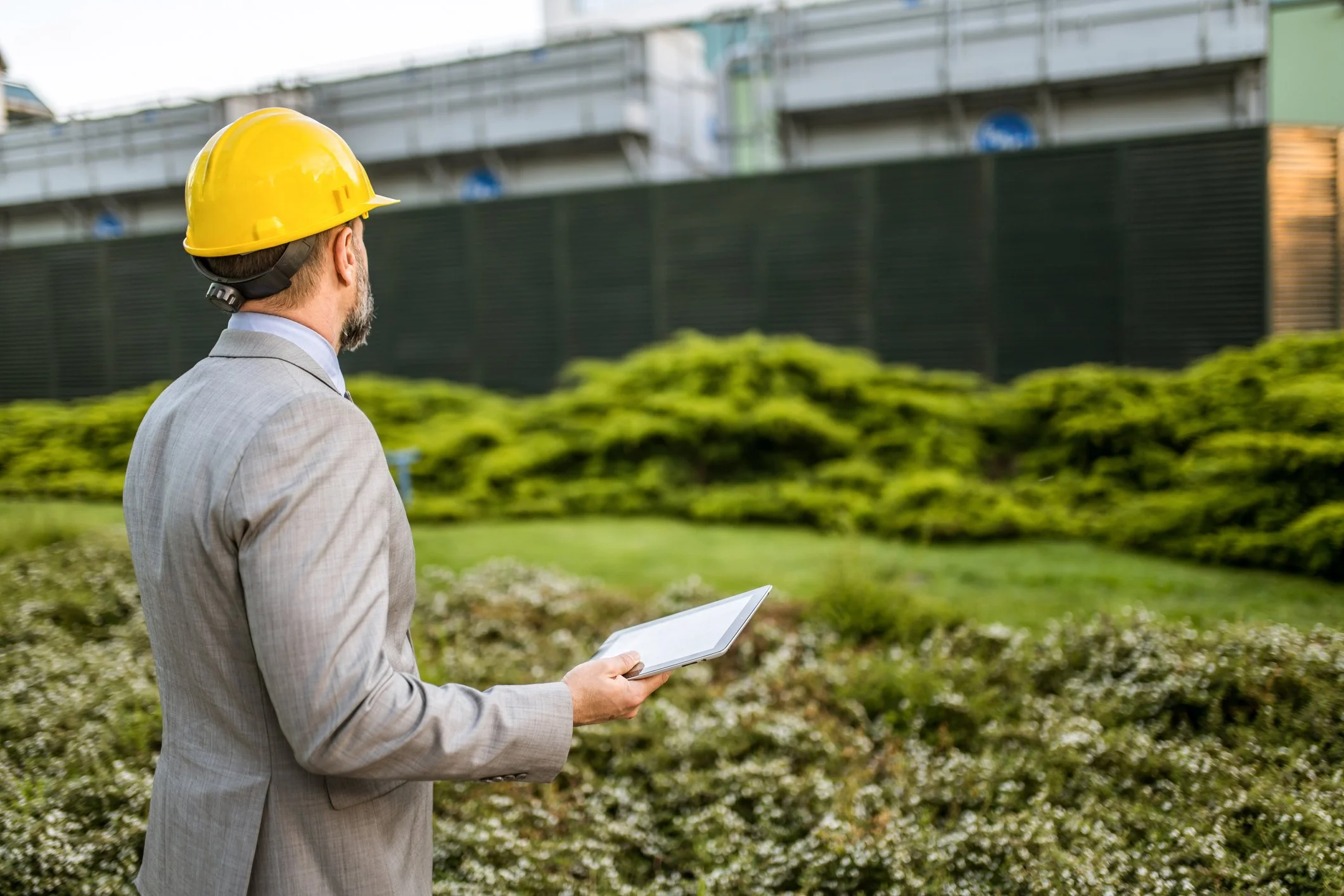 A man in a gray suit and yellow hard hat holding a tablet while standing outside near green bushes and a construction site.