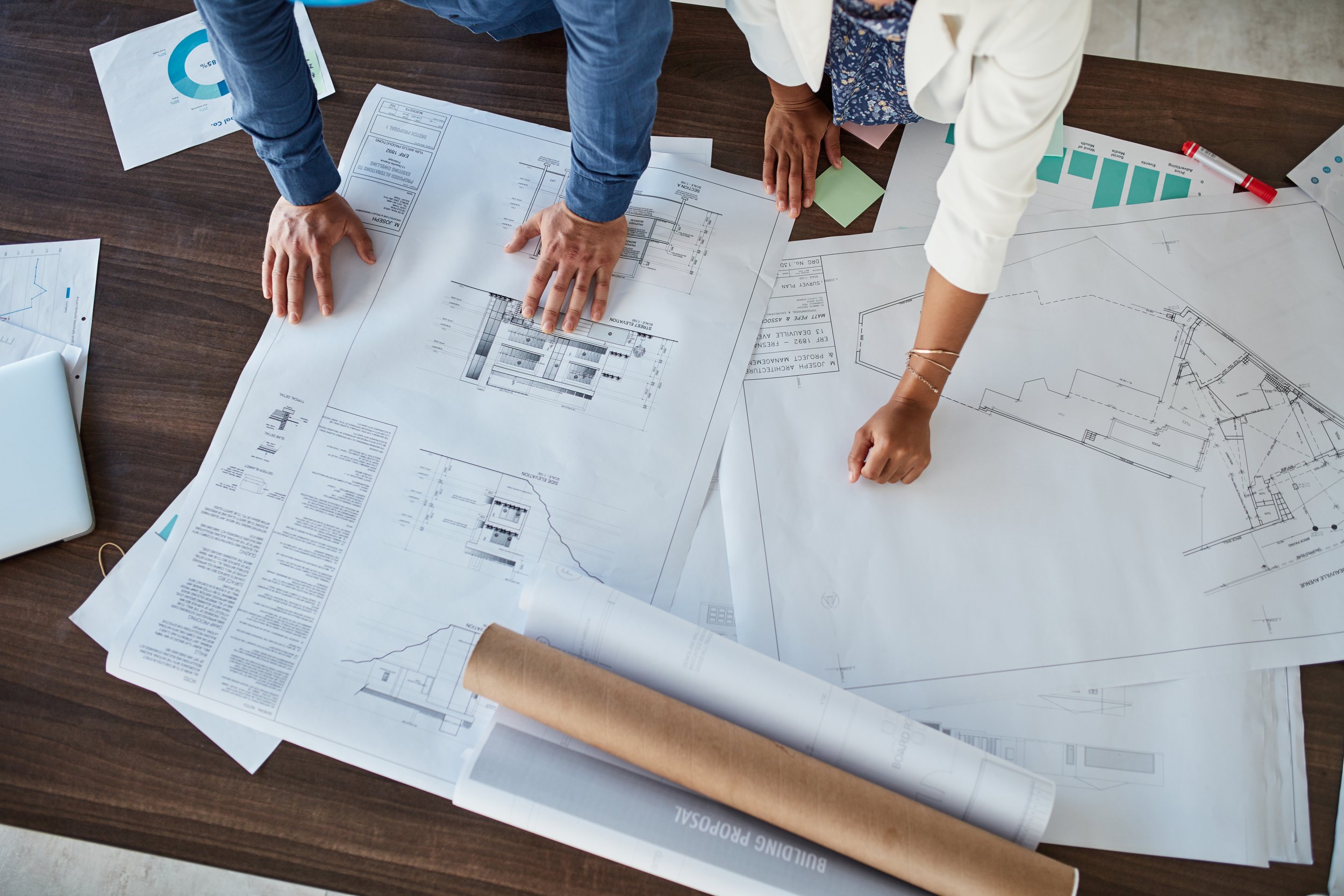 Two people reviewing architectural blueprints on a large wooden table. The table is also covered with rolled drawings, a pen, a laptop, and various papers.