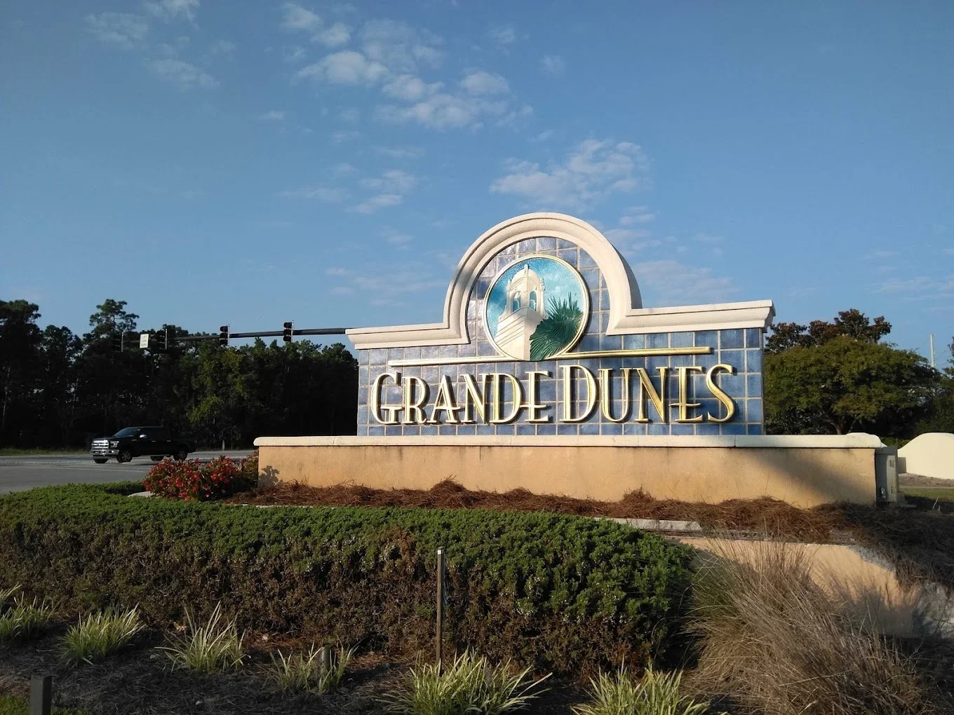 Entrance sign for Grand Dunes with blue and white design, featuring a lighthouse and palm tree, on a landscaped area under a clear sky.
