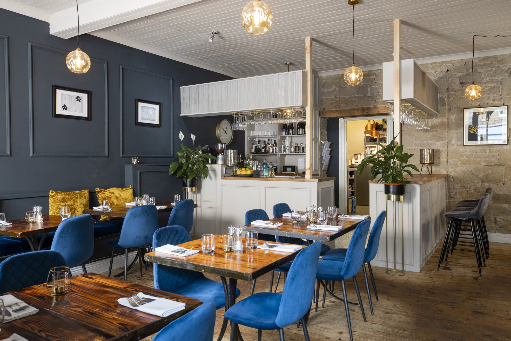Interior of a modern restaurant with wooden tables, blue velvet chairs, and a bar area with glassware and bottles, decorated with framed pictures, plants, and warm lighting.
