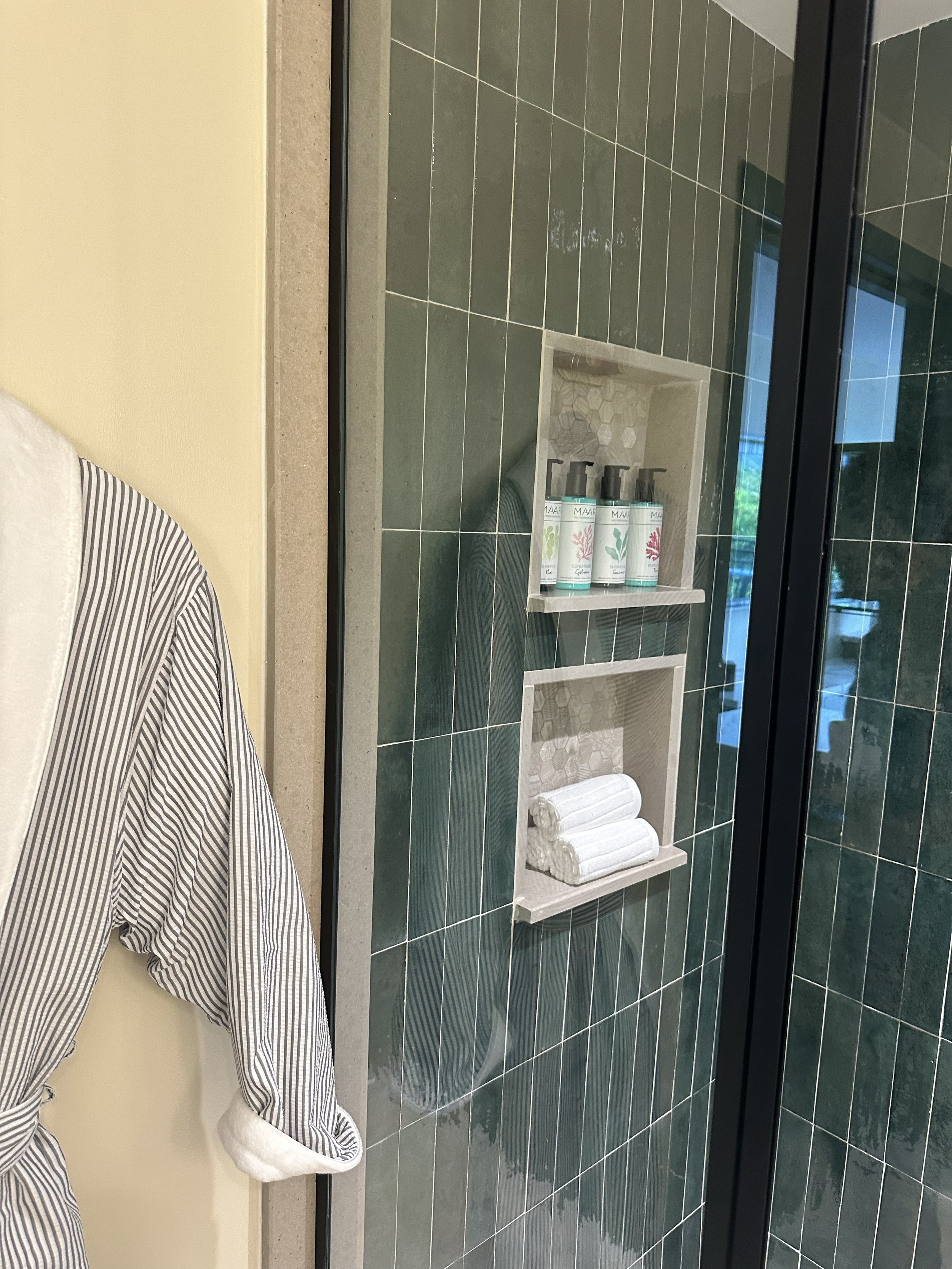 Shower area with green tiled wall, built-in shelf holding shampoo and conditioner bottles, and neatly folded white towels.