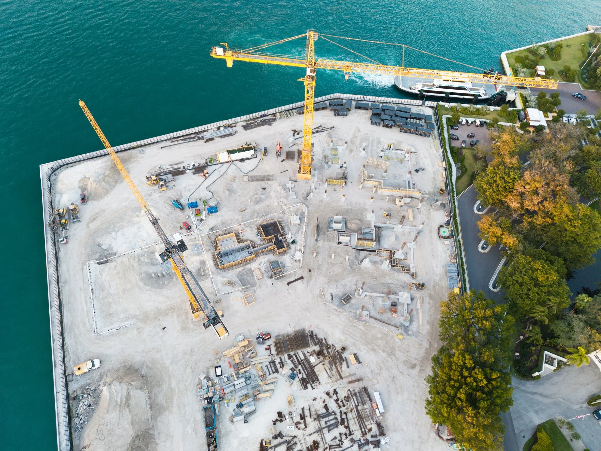 Aerial view of a construction site on a waterfront, with two large yellow cranes, construction materials, and partially built structures, adjacent to a parking lot and landscaped area with trees.