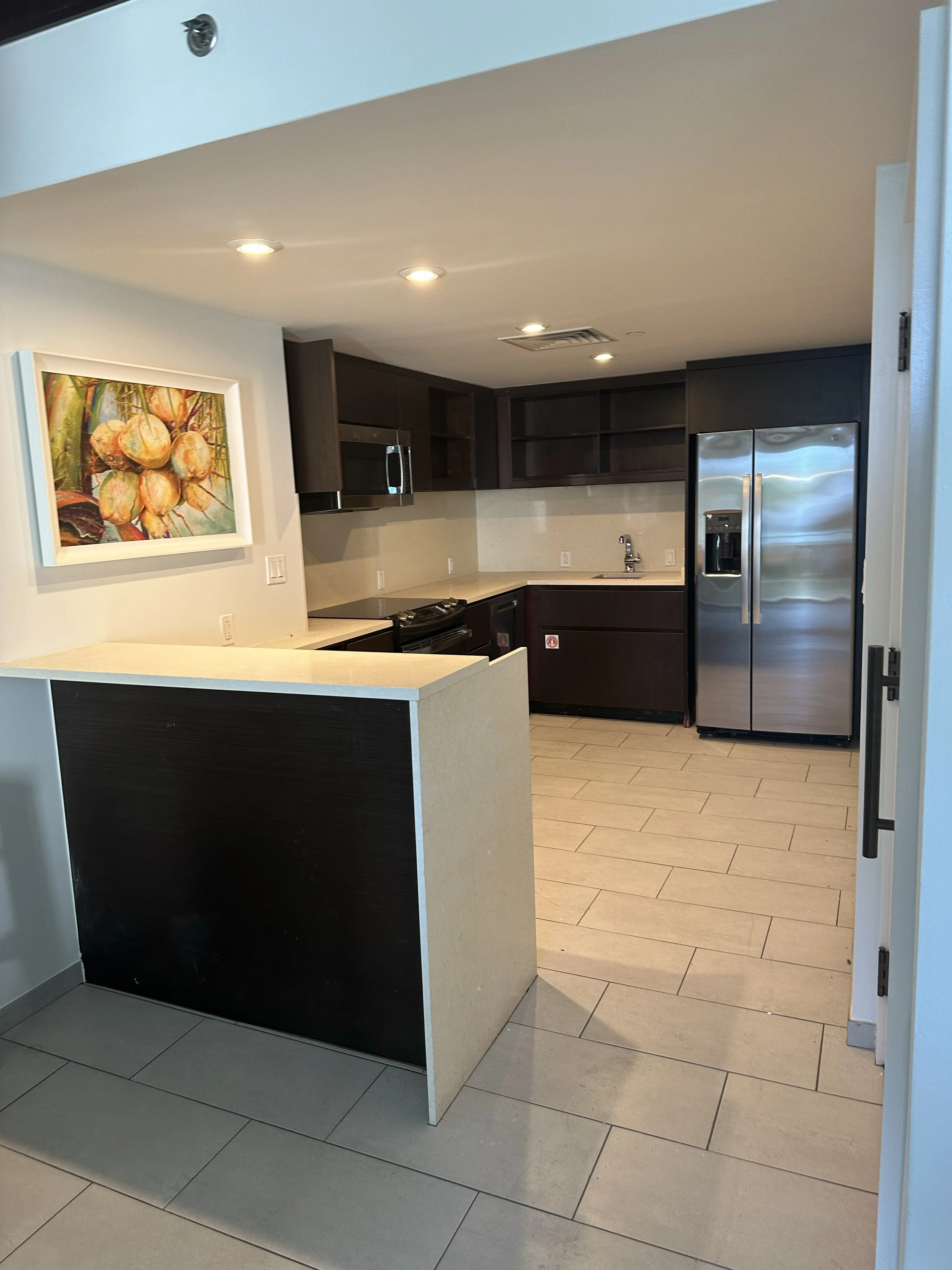 Modern kitchen with dark wood cabinets, stainless steel refrigerator, microwave, and oven. Beige countertops, beige tile flooring, and a colorful fruit-themed artwork on the light-colored wall.