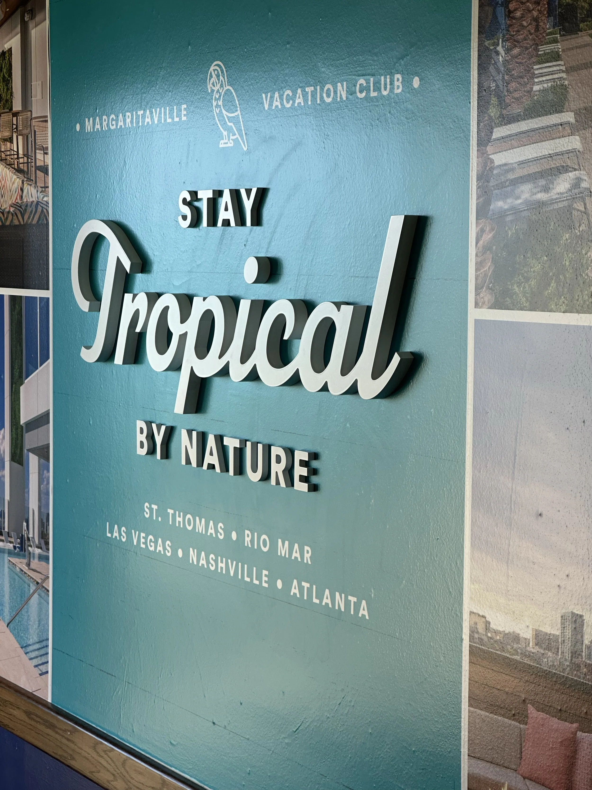 Sign with the message 'Stay Tropical by Nature' and locations listed: St. Thomas, Rio Mar, Las Vegas, Nashville, Atlanta, featuring a blue background and a small toucan illustration.