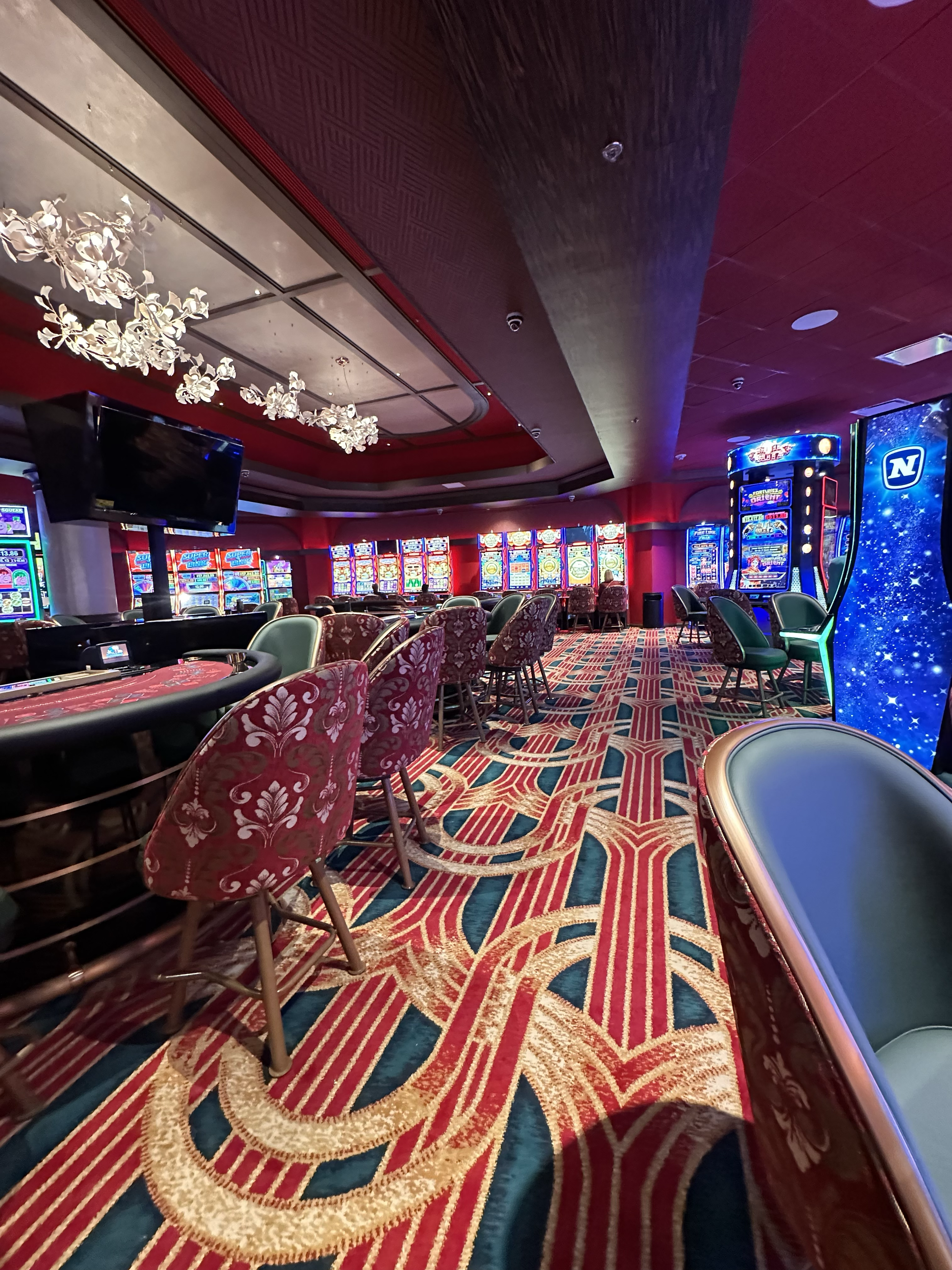 Inside a casino, with slot machines along the walls and a patterned carpet on the floor. There are chairs around tables and a television mounted on the wall, with decorative lighting on the ceiling.