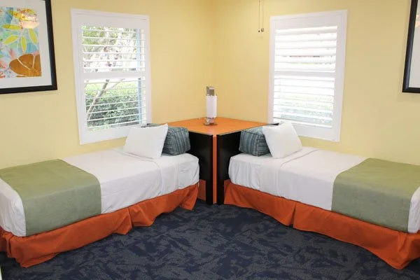 A bedroom with two twin beds with white pillows and green and orange bedding, separated by a corner table with a lamp, yellow walls, two windows with white blinds, and framed artwork on the walls.
