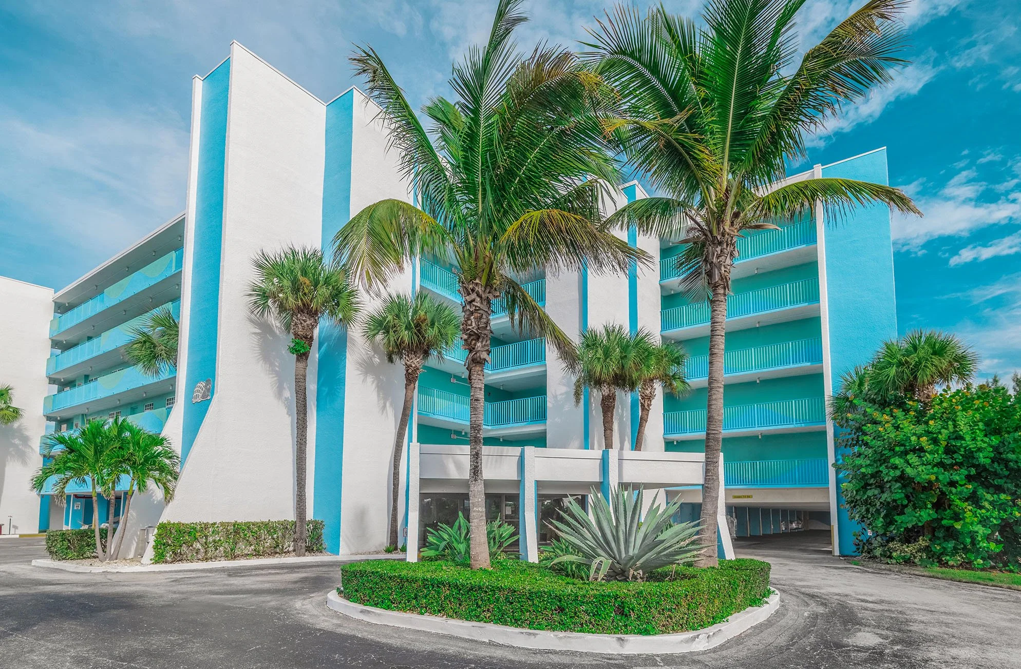 Multi-story hotel building with blue accents, surrounded by palm trees and greenery under blue sky with clouds.