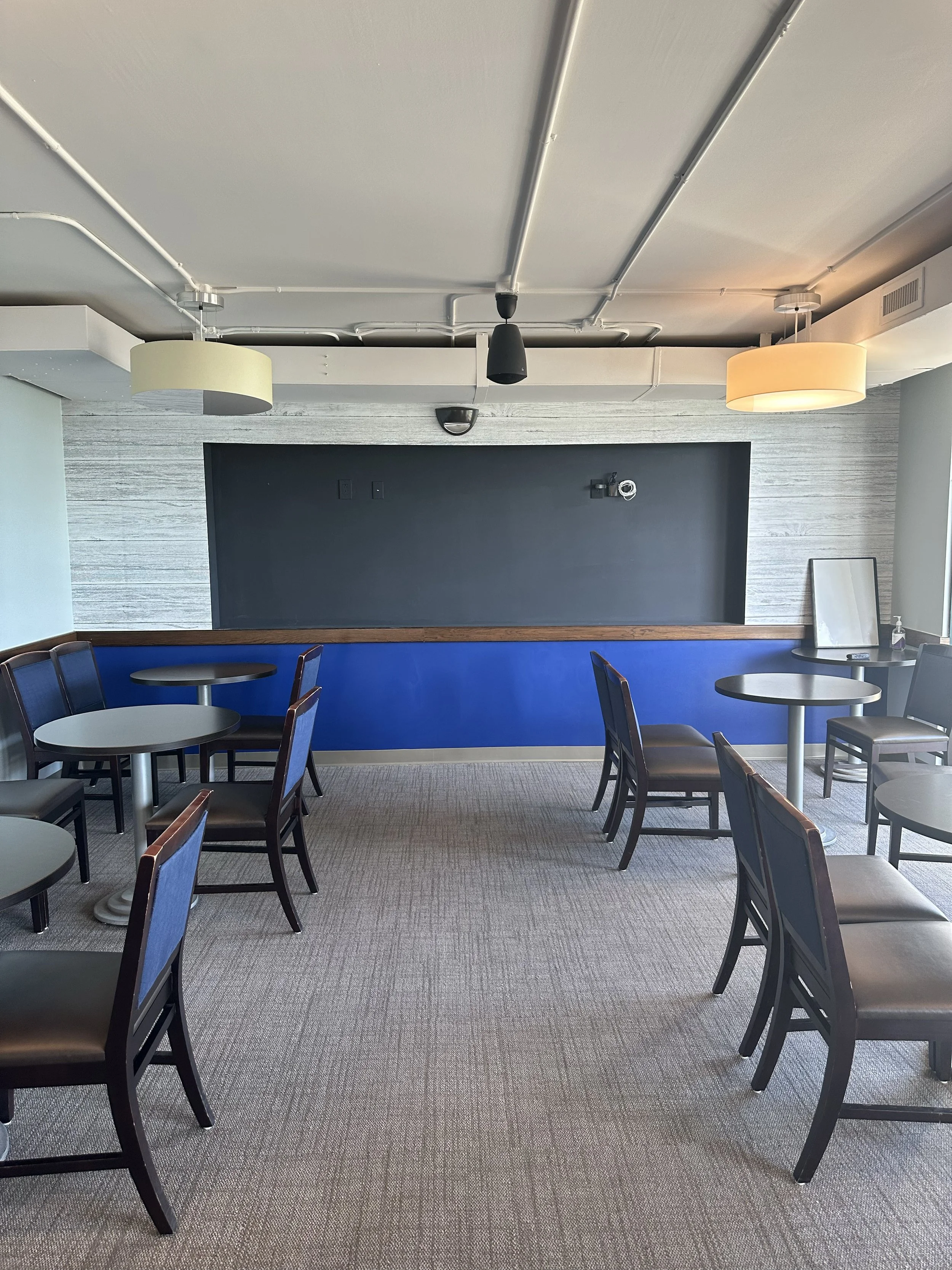 Empty indoor dining area with round tables and chairs, black chalkboard wall, and modern lighting fixtures.