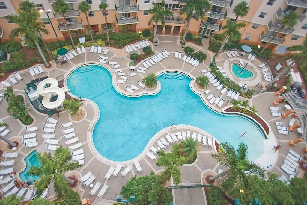 An aerial view of a hotel pool area with a large, irregularly shaped swimming pool, surrounded by numerous lounge chairs and palm trees. There are also smaller pools and a shaded seating area nearby.