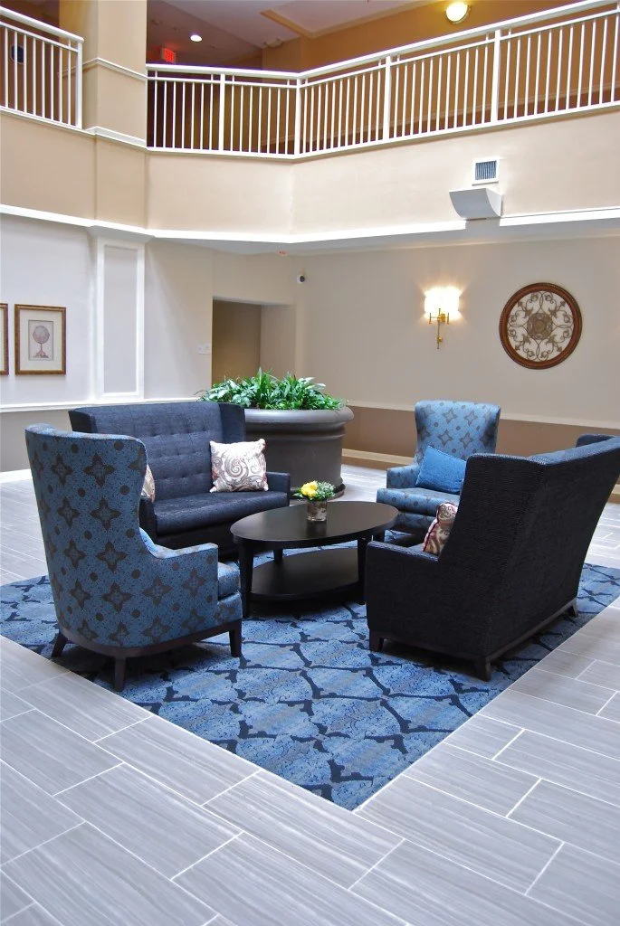 A cozy sitting area with four blue and black upholstered armchairs arranged around a round black coffee table, positioned on a blue patterned rug in an indoor lobby or common area of a building.