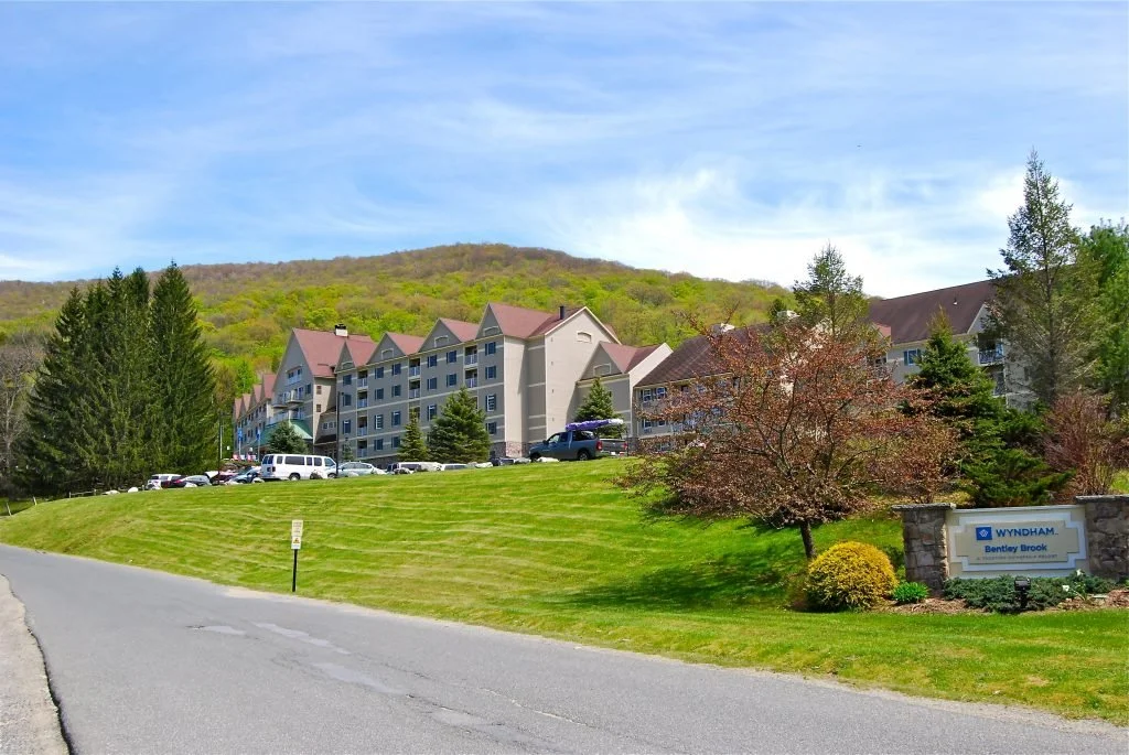 Residential complex with multiple buildings, cars parked outside, surrounded by trees and greenery, on a hillside with a mountain in the background, under a partly cloudy sky.