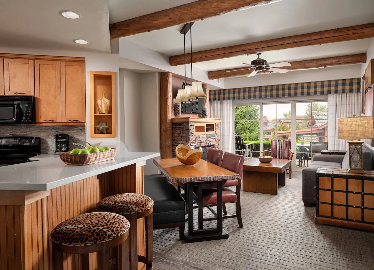 Living room and kitchen area with wooden beams, sliding glass door with curtains, and a view of outdoor greenery.