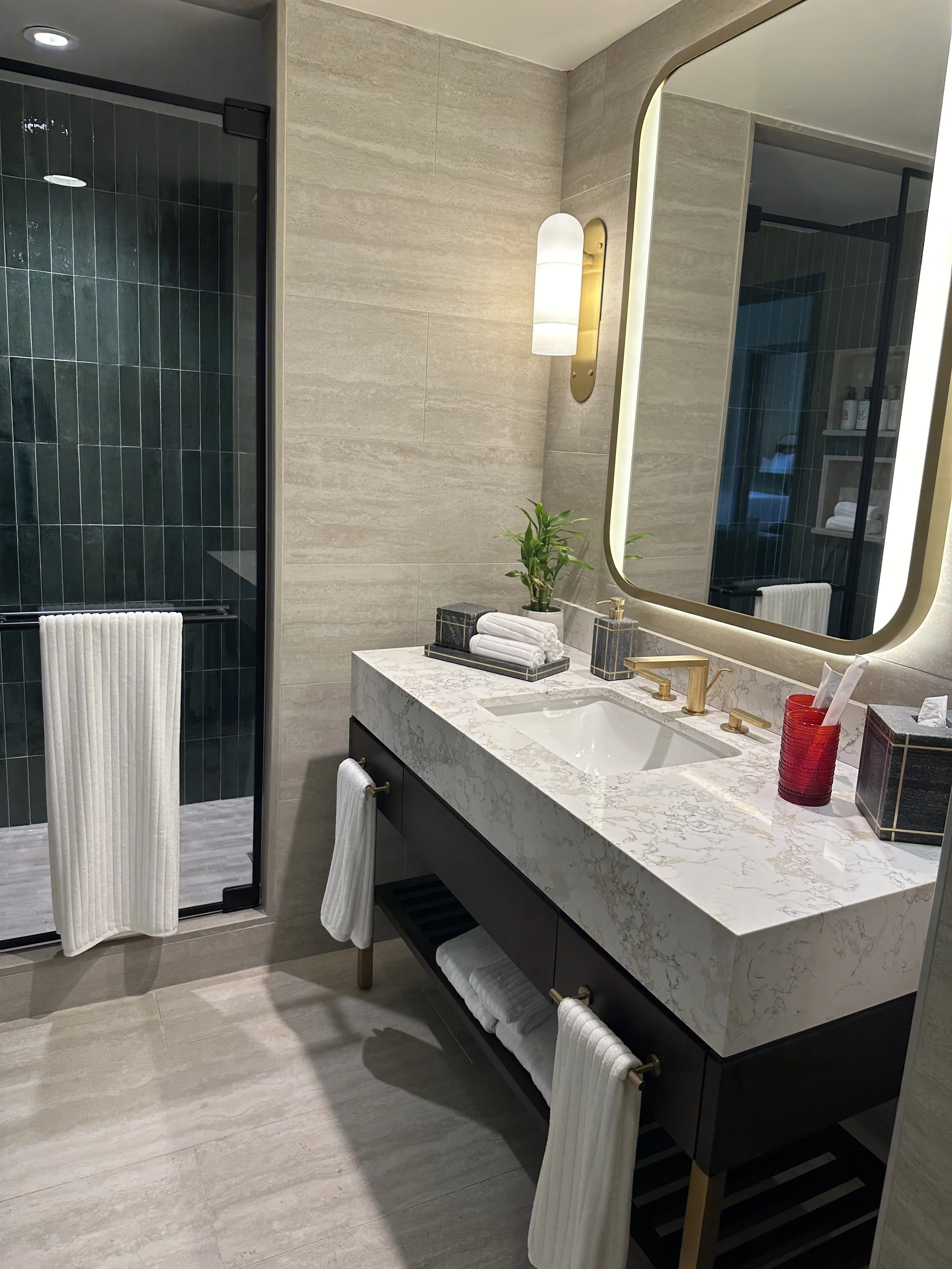 Modern bathroom with light beige tiled walls and flooring, a marble countertop sink, gold fixtures, a large illuminated mirror, and a walking shower with dark green tiles and a white towel hanging on the shower door.