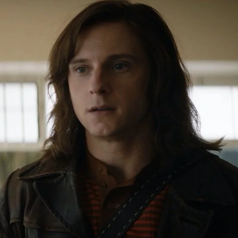 A man with shoulder-length brown hair and blue eyes, wearing a black leather jacket over a red and black checkered shirt, standing indoors with a neutral expression.