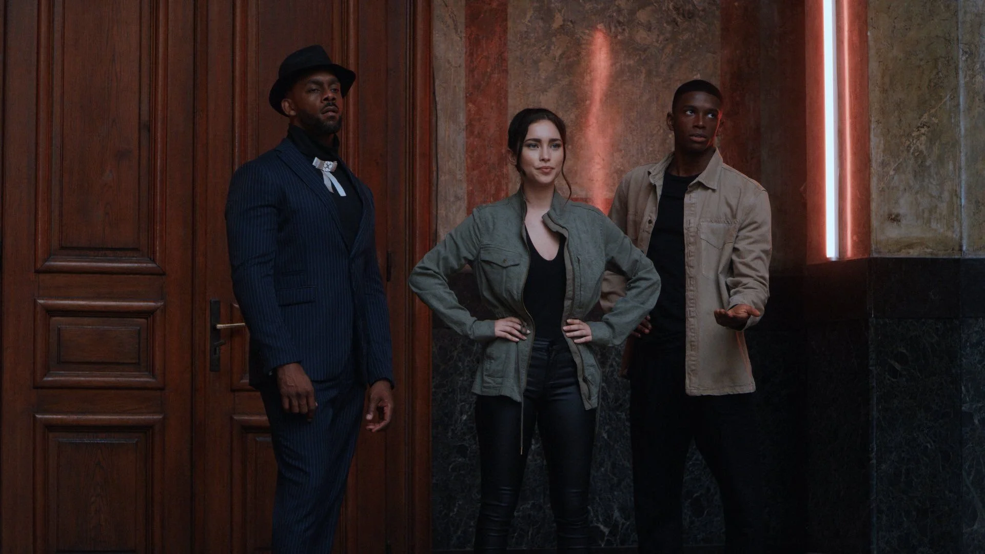 Three young adults standing in front of a wooden door and marble wall, posing for a photo. The man on the left is wearing a dark pinstripe suit, black shirt, and black hat. The woman in the middle has dark hair, wearing a green jacket and black pants. The man on the right is wearing a beige jacket over a black shirt, with his right hand extended gesturing to the side.
