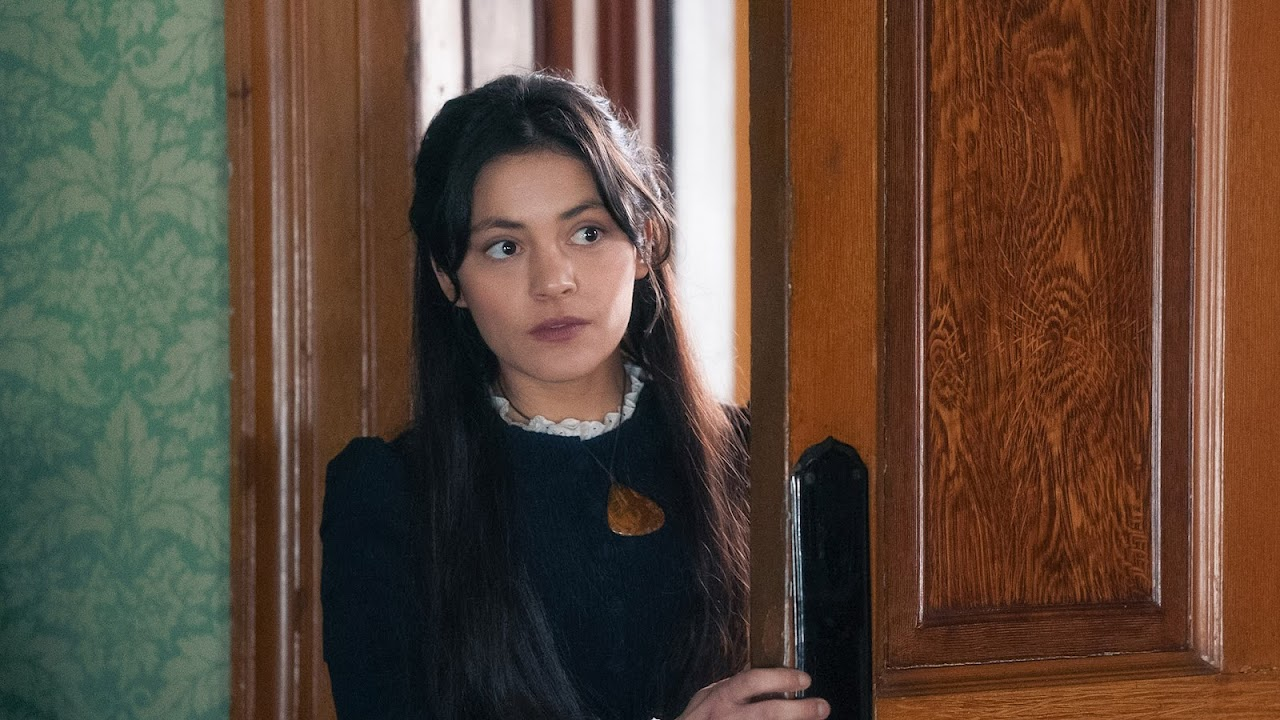A young woman with long dark hair looking through a partially opened wooden door in a vintage-style room.