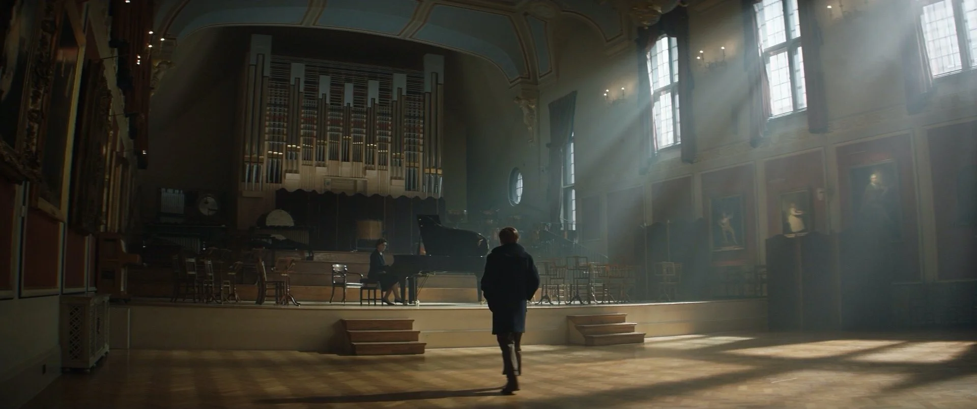 A large, elegant concert hall filled with soft natural light streaming in through tall windows. In the center, a grand piano is being played by a person, with another person sitting nearby. The stage is set with chairs and musical instruments, and the background features a large pipe organ and portraits on the walls.