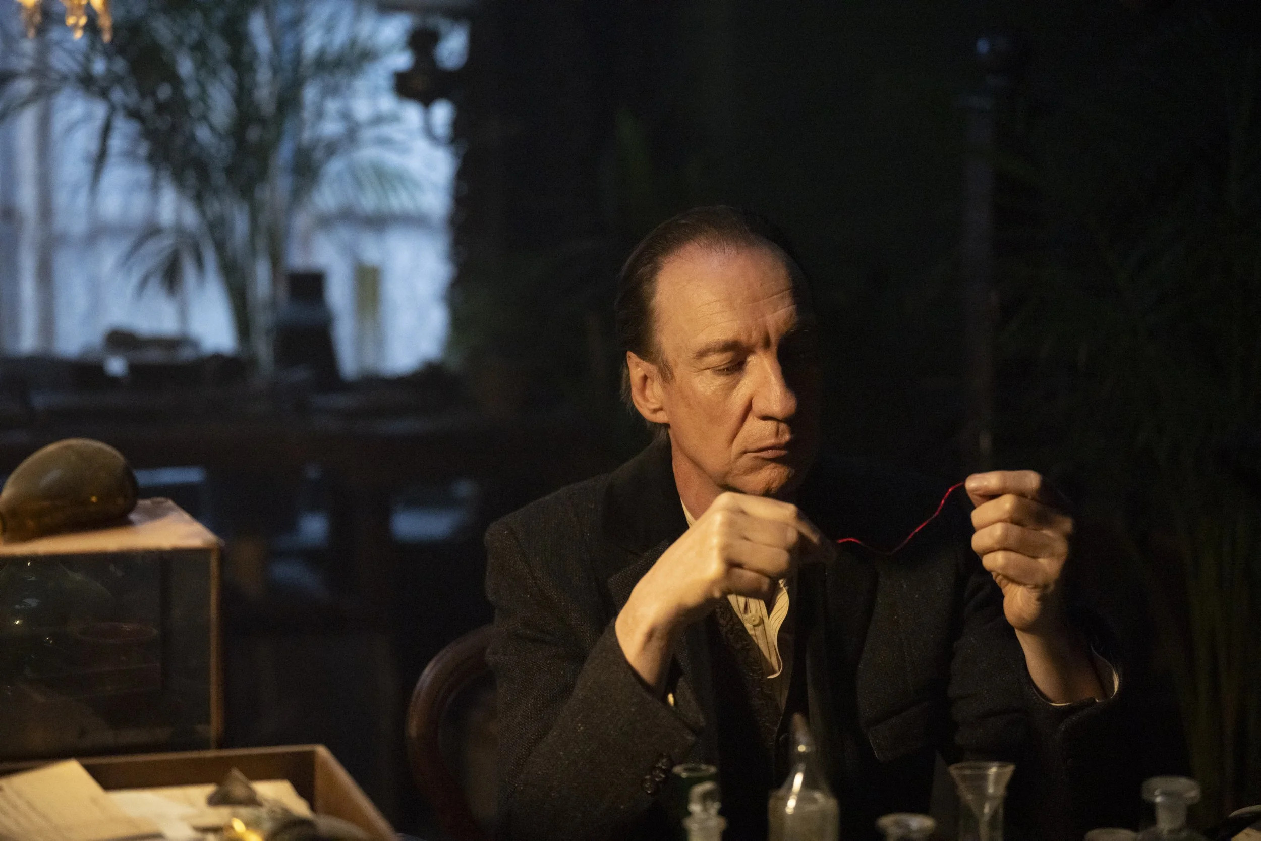 A man with slicked-back hair and wearing a dark blazer is focused on a task involving red string or wire. The scene looks like it is set indoors at night, with a dark background and some lighting highlighting the man's face and hands.