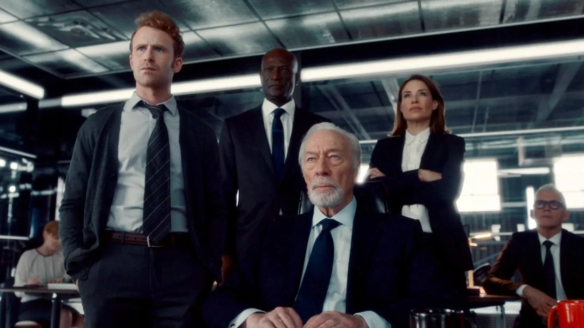 Four people in formal business attire standing and sitting in an airport terminal with their arms crossed or resting, looking serious or focused.