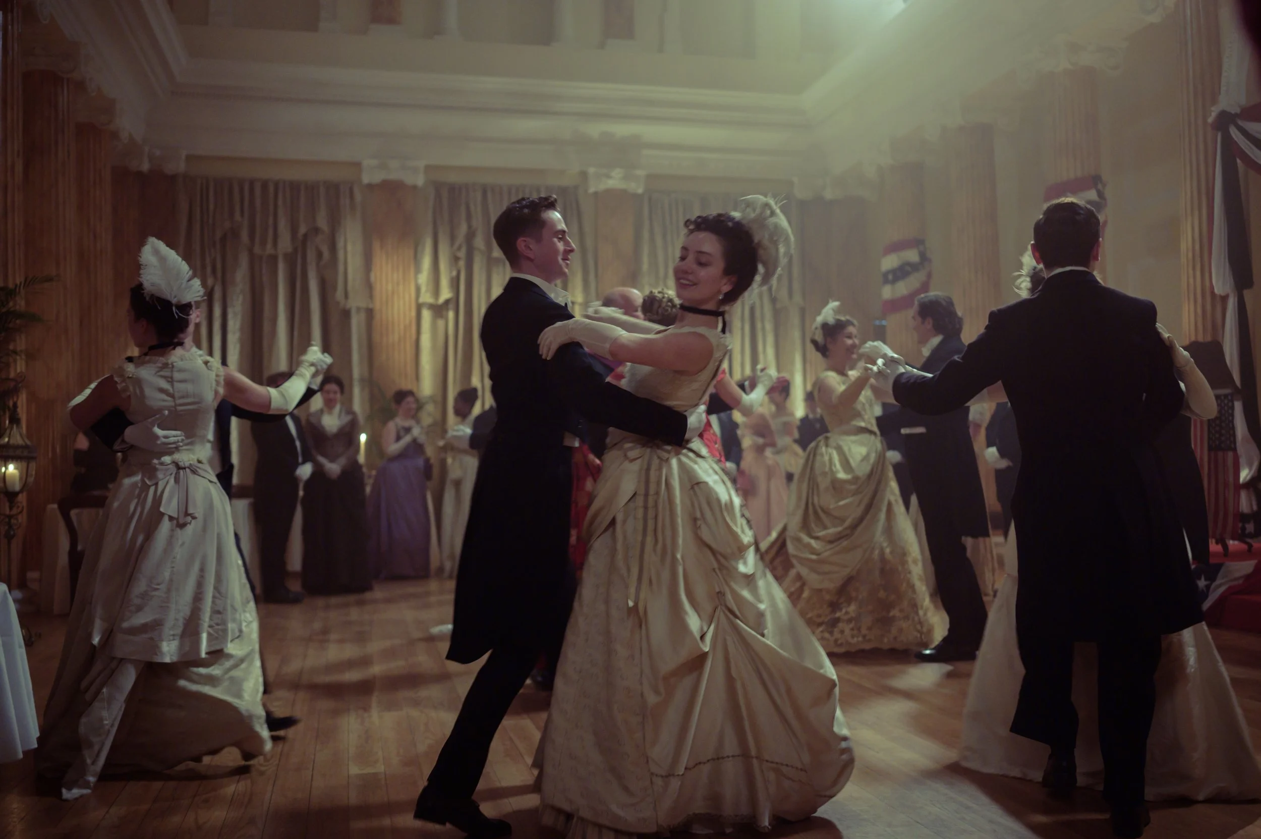 Couple dancing at a ballroom dance event in period costumes, surrounded by other dancers in similar attire, in an elegant, wood-paneled room.