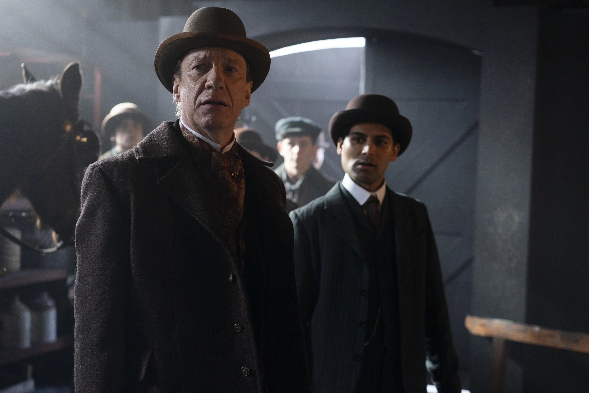 A scene from a period film or show featuring two men in vintage clothing and bowler hats, with other characters and horses in the background, in a historical setting.