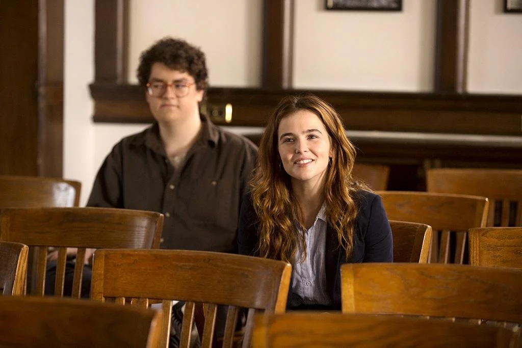 A woman with long, wavy brown hair smiling in a courtroom or conference room with wooden chairs and paneling, accompanied by a man with curly hair and glasses in the background.