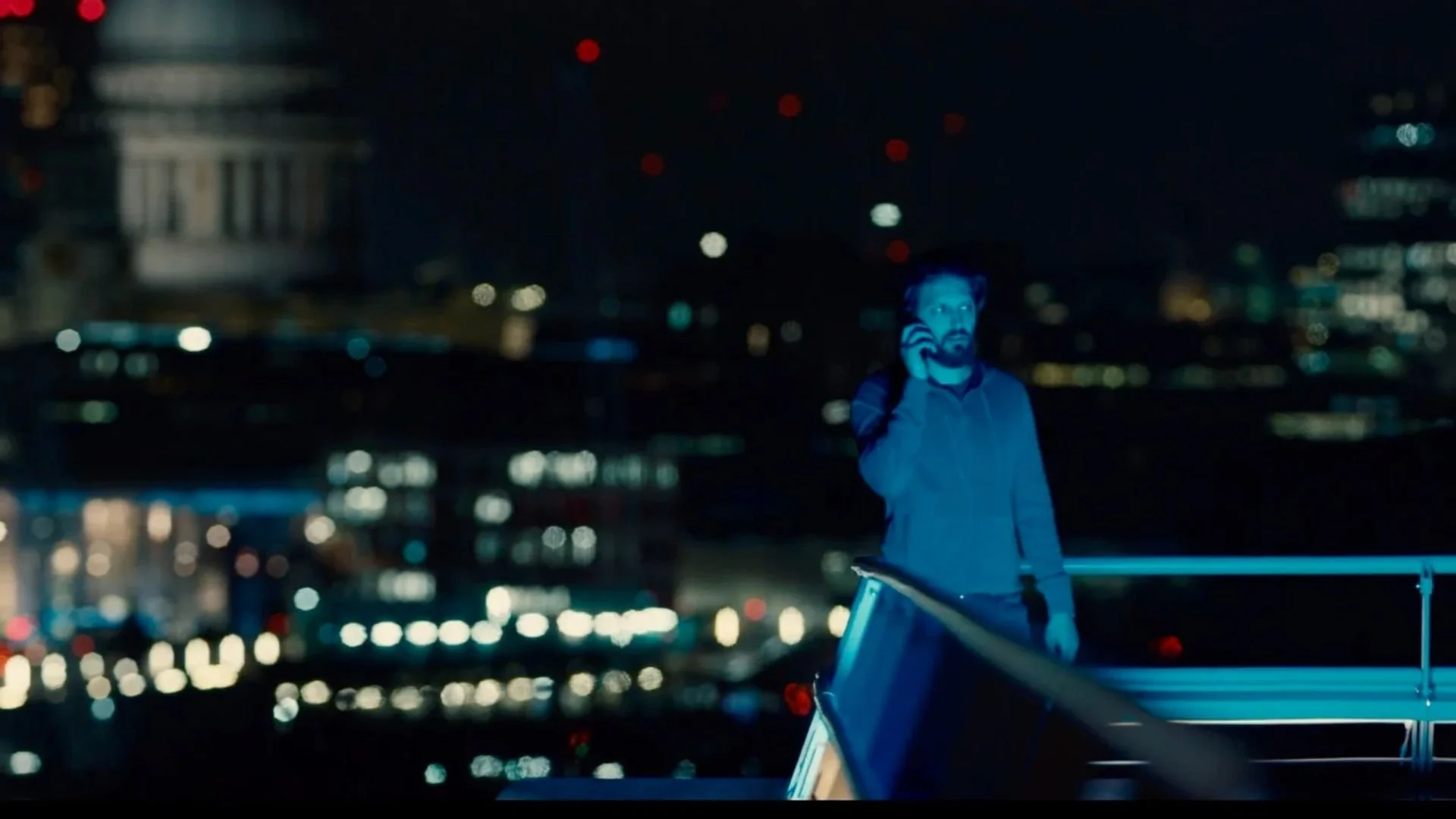 A man standing on the roof of a vehicle at night, talking on his cellphone, with city buildings and lights blurred in the background.