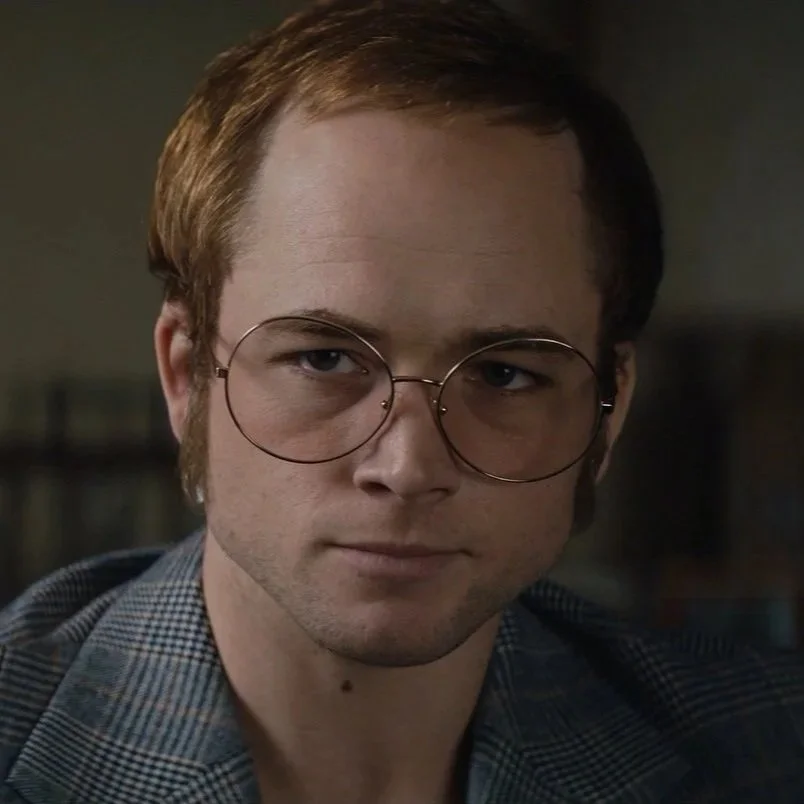 Close-up of a young man with short brown hair, wearing large round glasses and a plaid shirt in a dimly lit room.