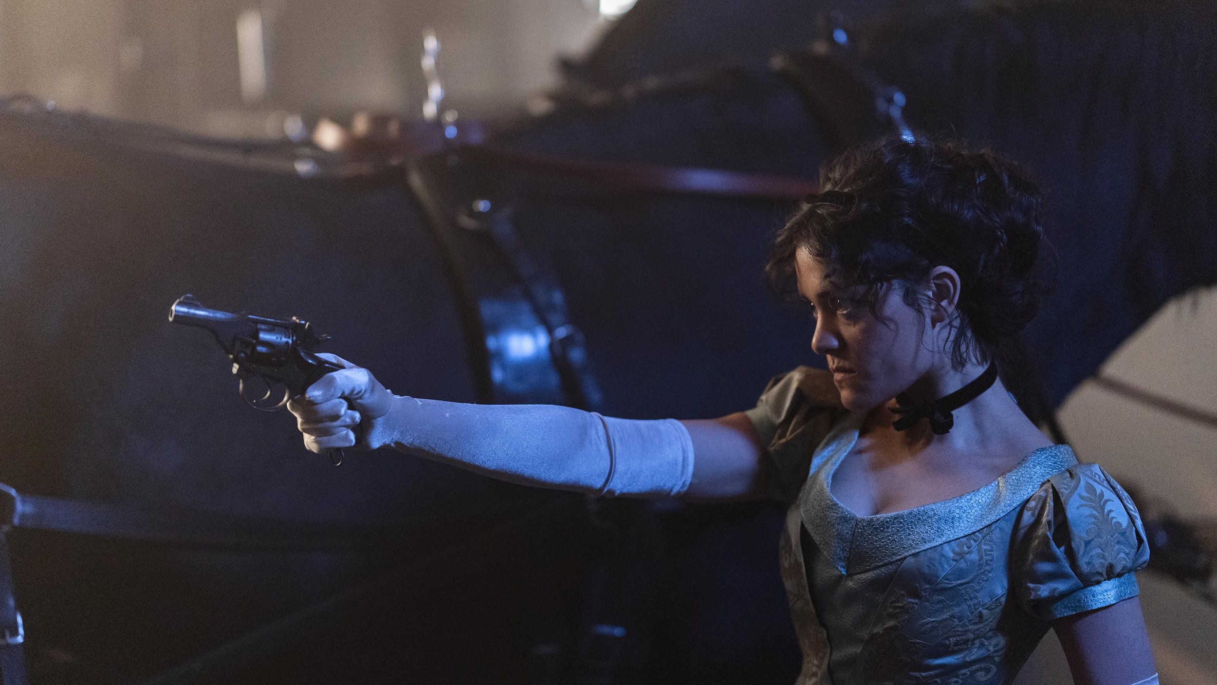 A woman with short curly hair, dressed in a vintage style dress and gloves, aiming a revolver in a dark room.