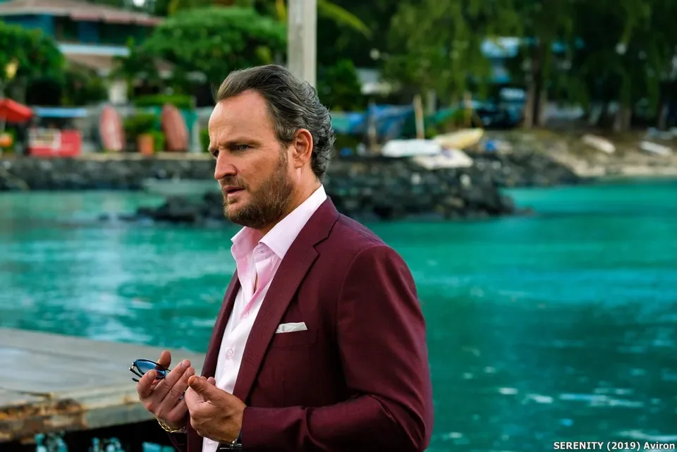 A man with dark hair and a beard wearing a maroon suit and pink shirt, standing outdoors near a body of water with boats and trees in the background.