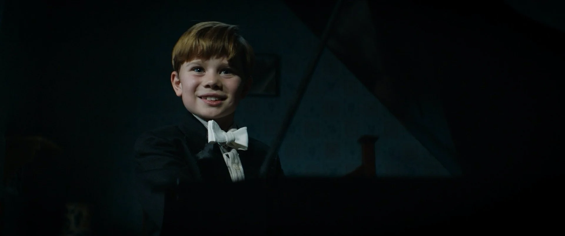 A young boy with red hair, wearing a tuxedo with a white bow tie, sits in a dark room and smiles.