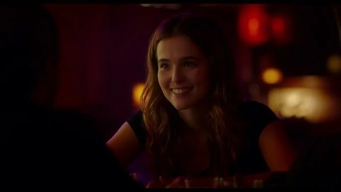 A young woman with brown hair smiling in a dimly lit setting, possibly a bar or restaurant.